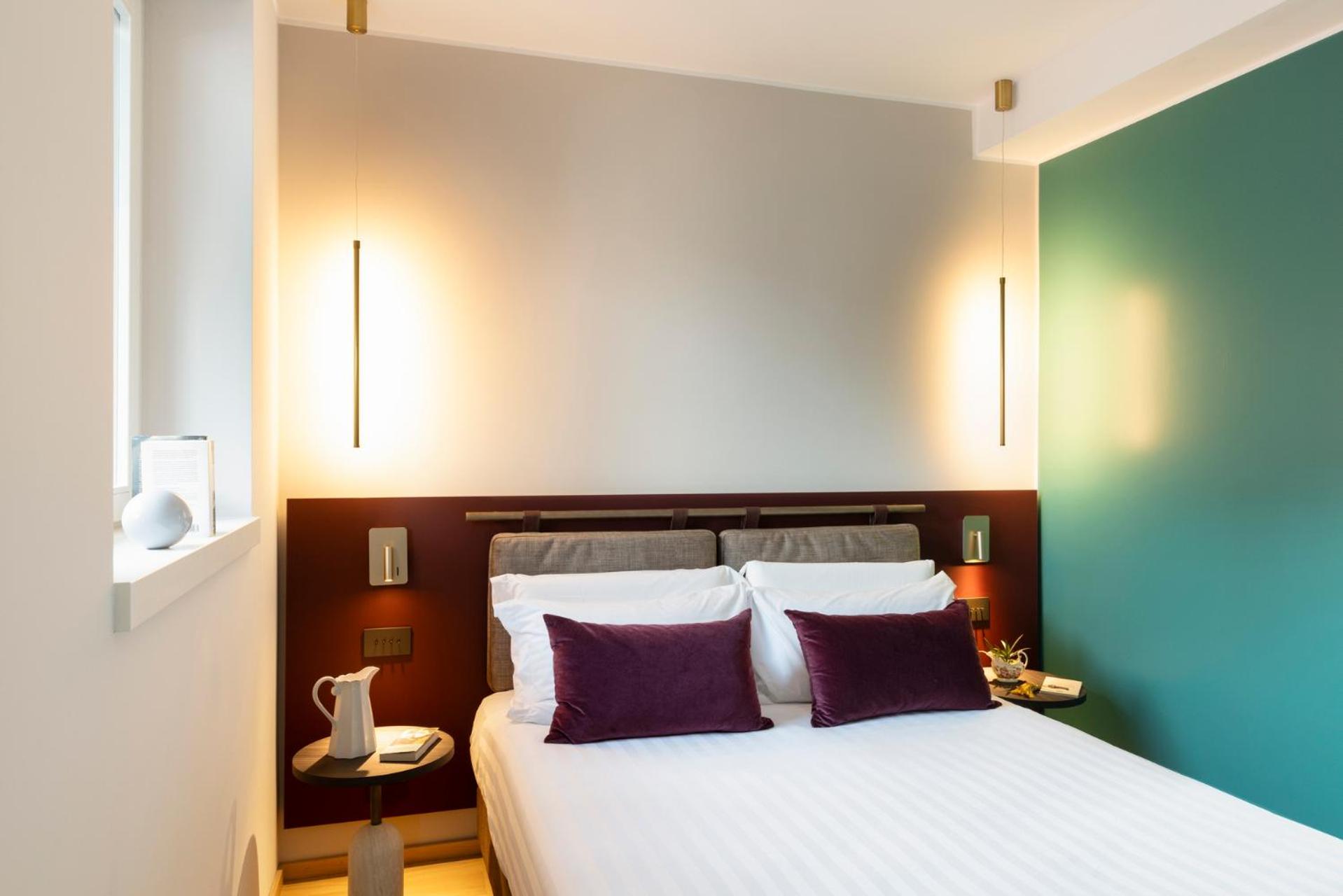 A bed flanked by hanging linear lights against a burgundy and cream wall beside a green partition.