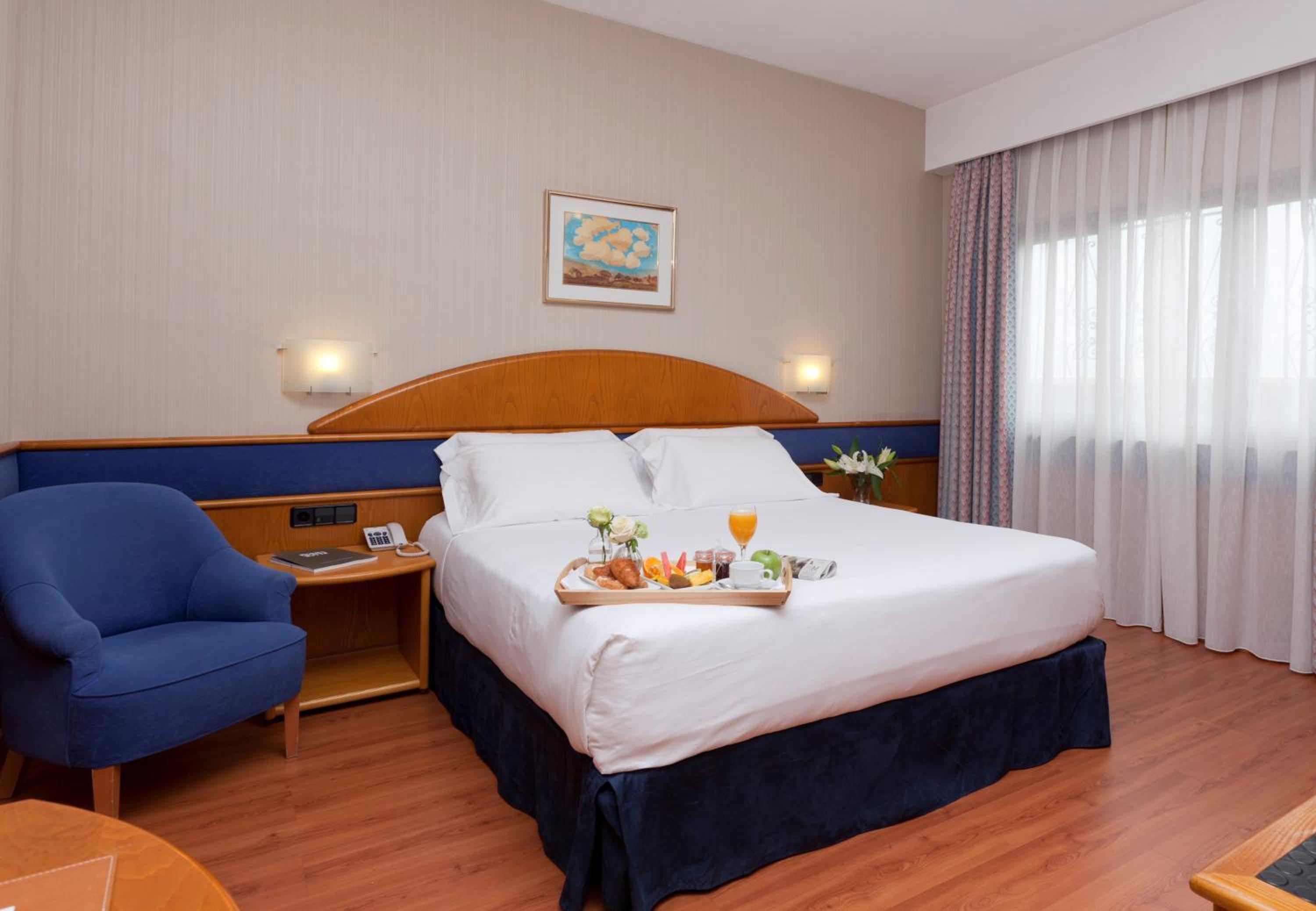 A hotel bedroom with a bed holding a breakfast tray, wooden headboard, and a blue armchair.
