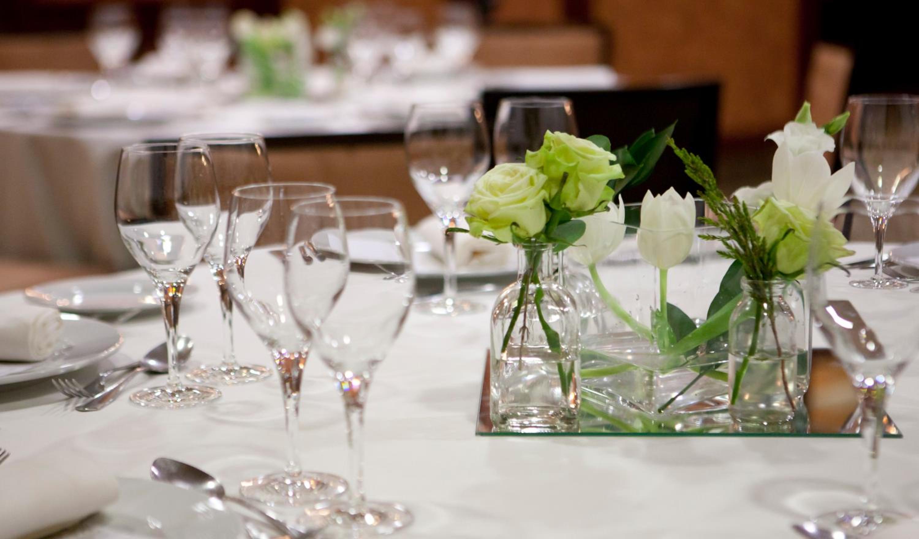 A banquet table set with stemmed wine glasses and pale green roses in small glass bottles on a white tablecloth.