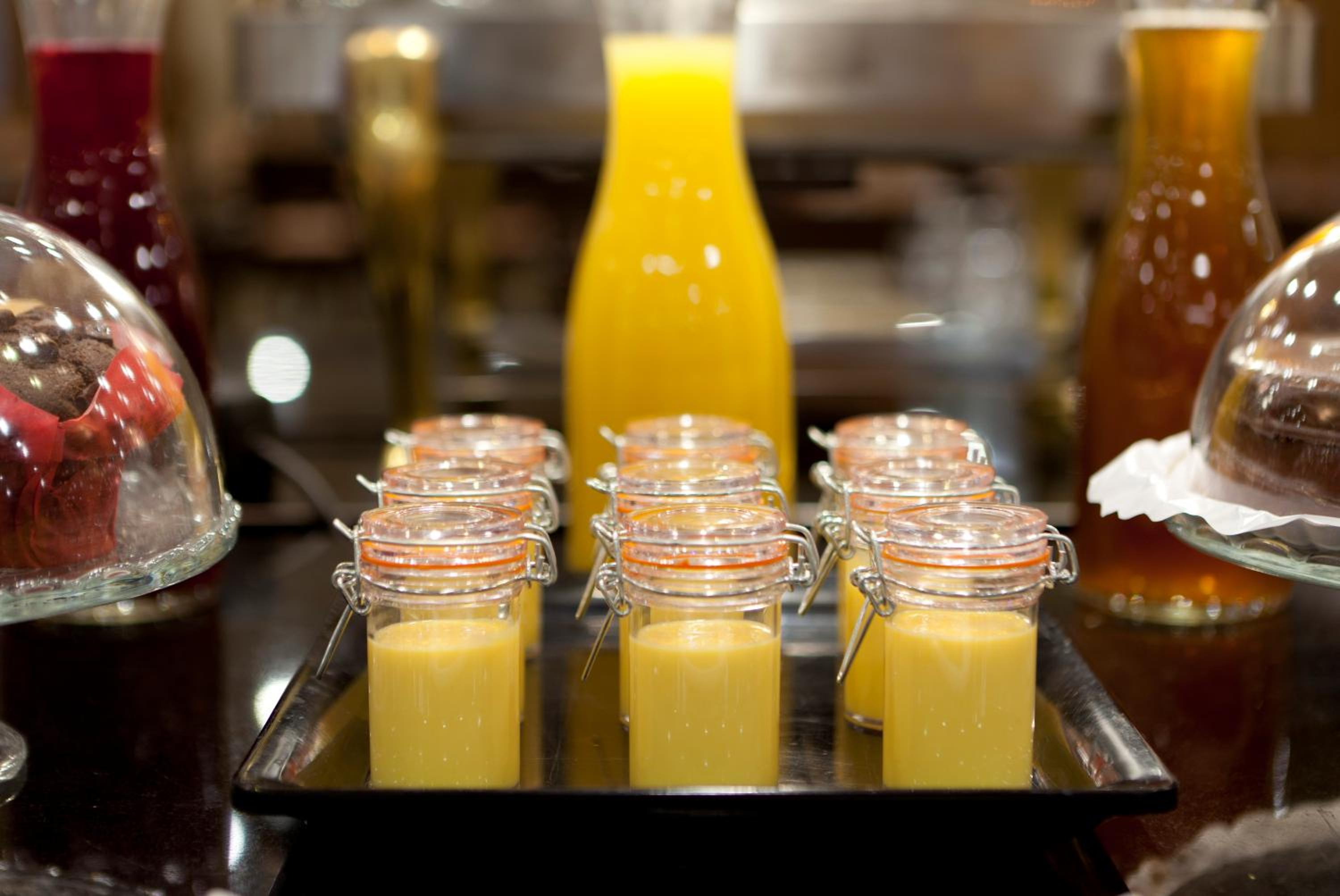 Glass jars filled with yellow mousse or smoothie on a buffet tray in front of a glass decanter of juice.