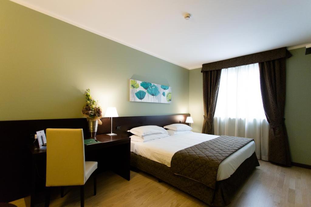 A hotel bedroom with green walls, light wood flooring, a bed, and dark drapery framing a sunny window.