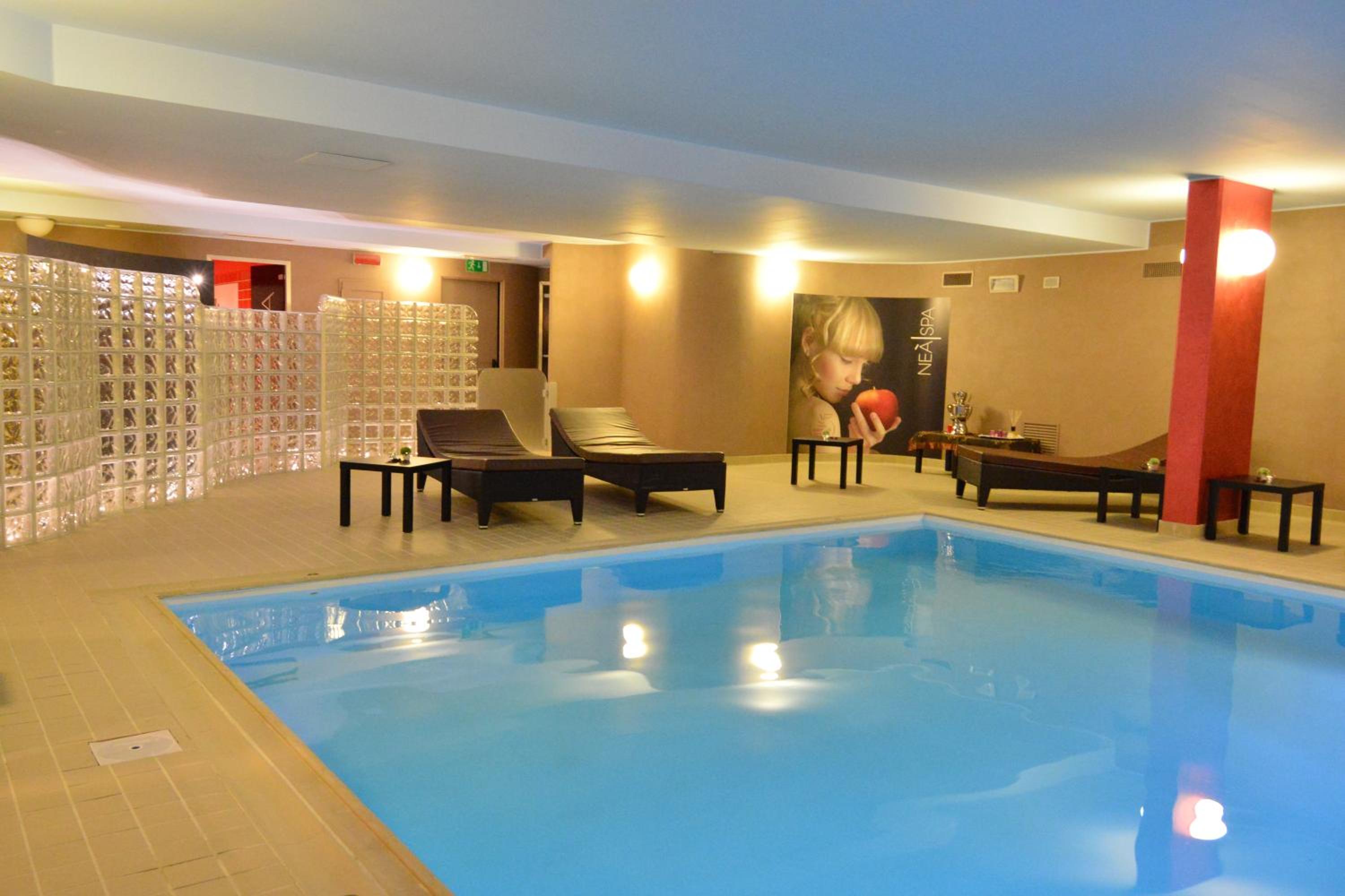 An indoor swimming pool with blue water, tiled surround, lounge chairs, and warm wall lighting.