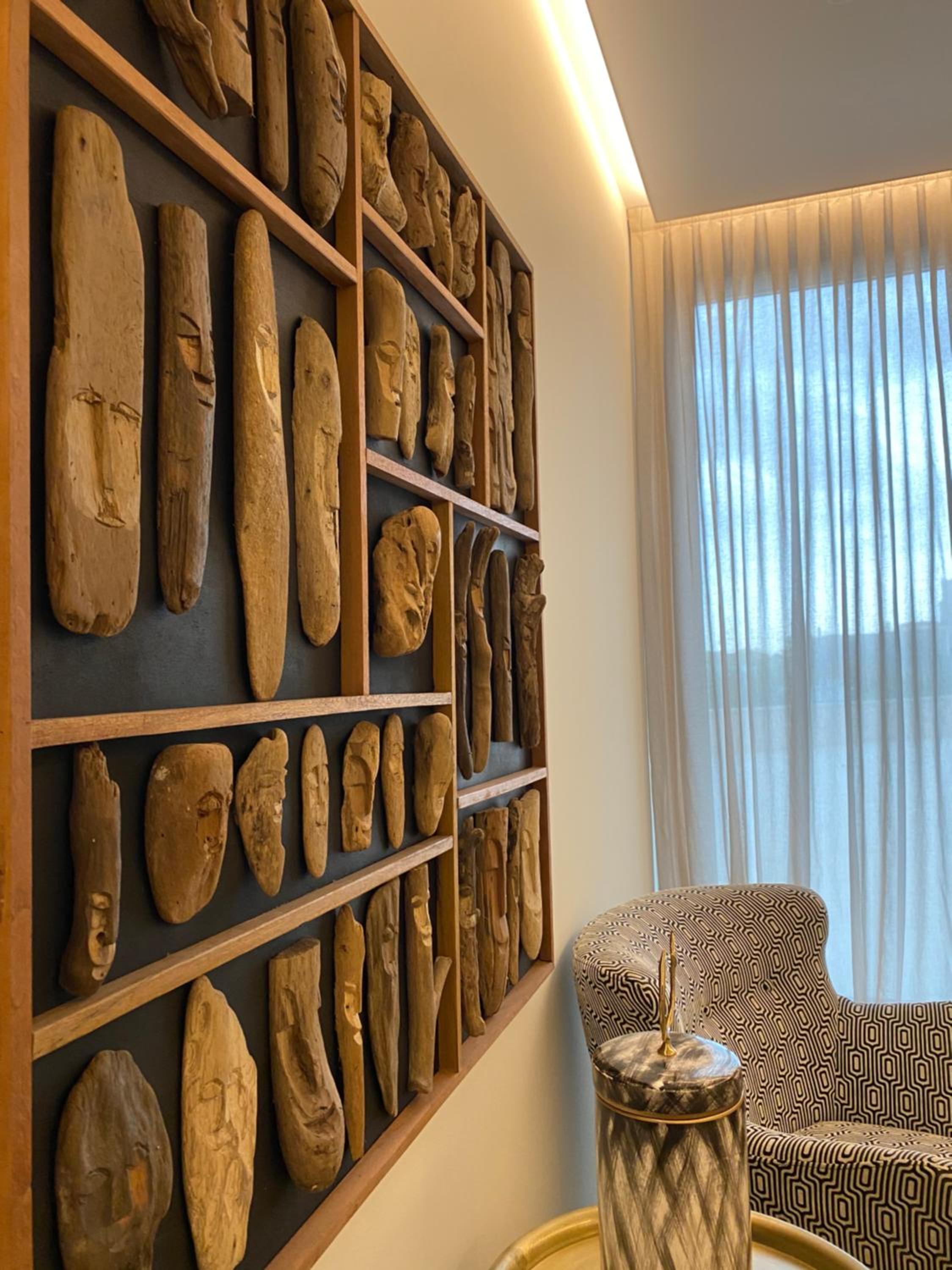 Framed carved driftwood faces hang on a wall beside a patterned armchair and sheer curtains.