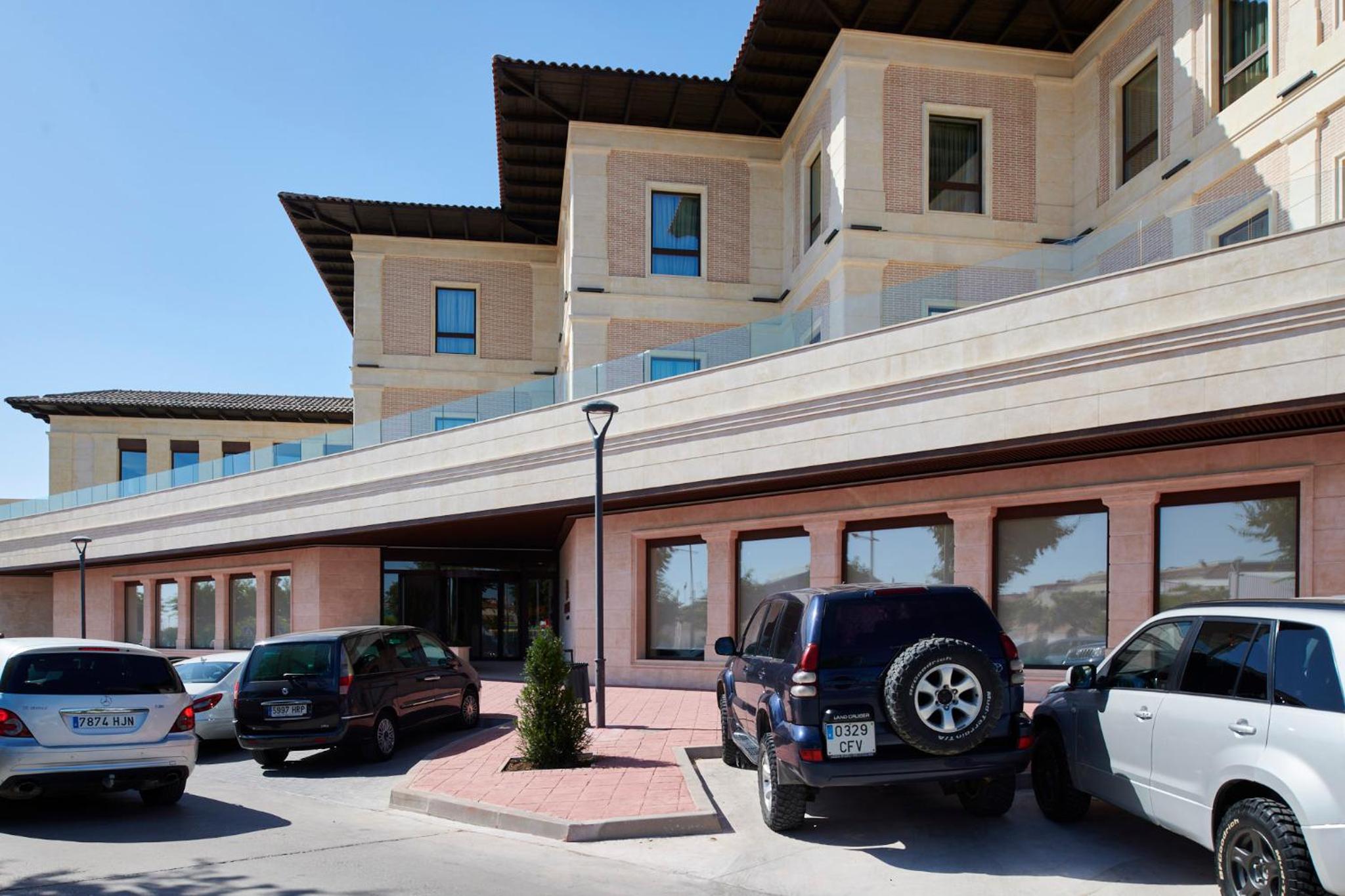 Parked cars sit in front of the stone facade of Ansares Hotel under clear daylight.