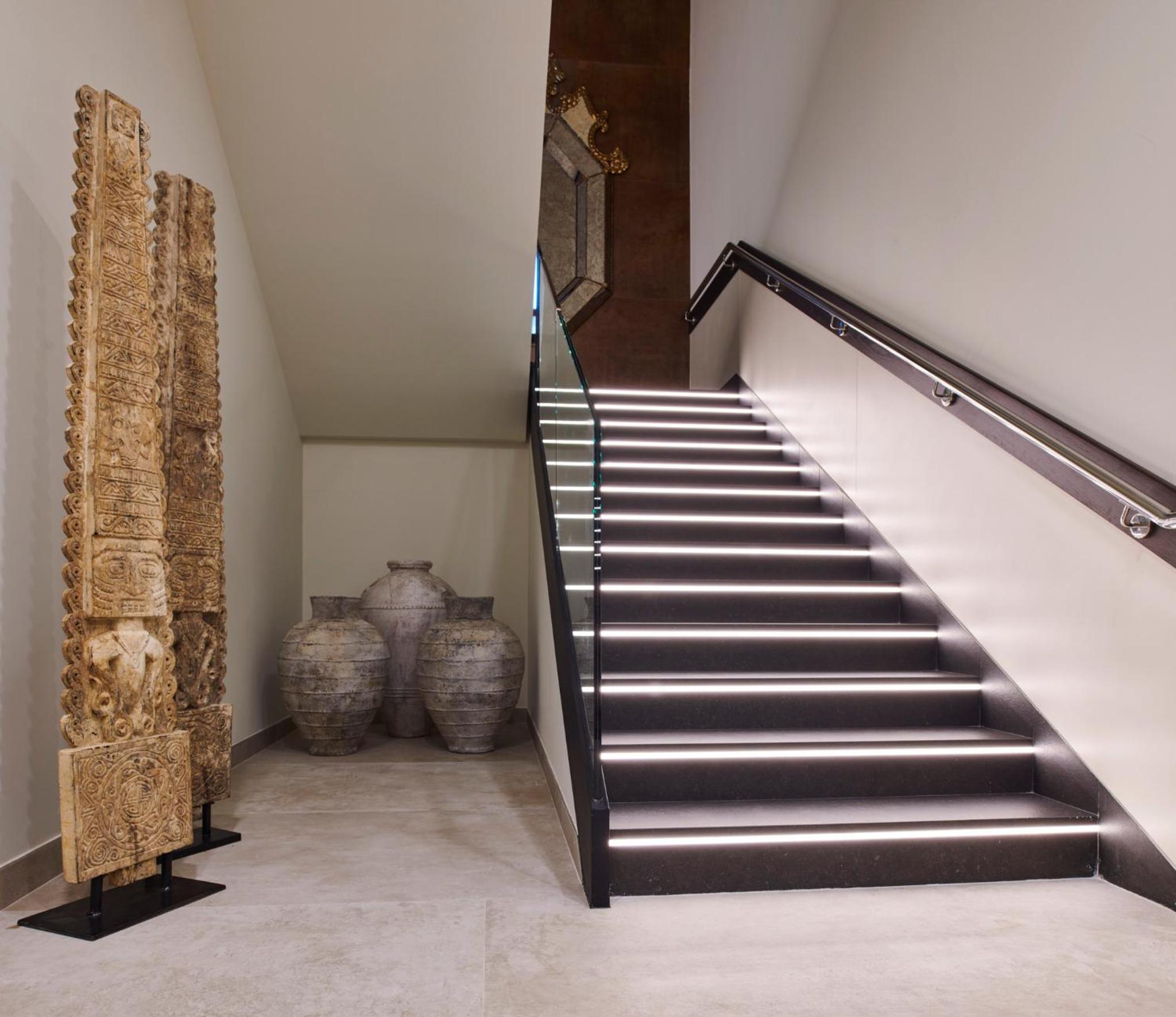Illuminated stairs with glass railings rise past tall carved stone columns and weathered clay urns.