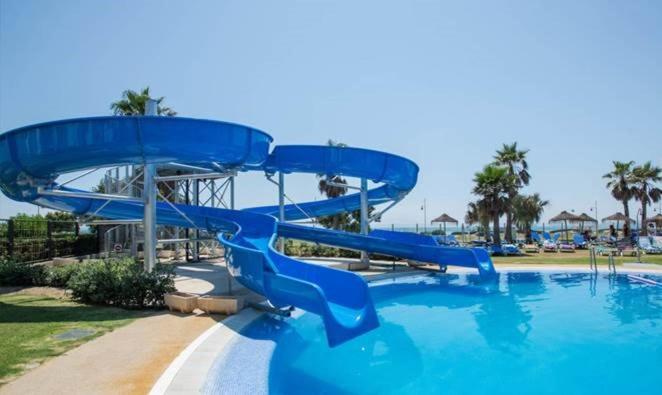 A blue spiral water slide curves into an outdoor swimming pool bordered by palm trees.
