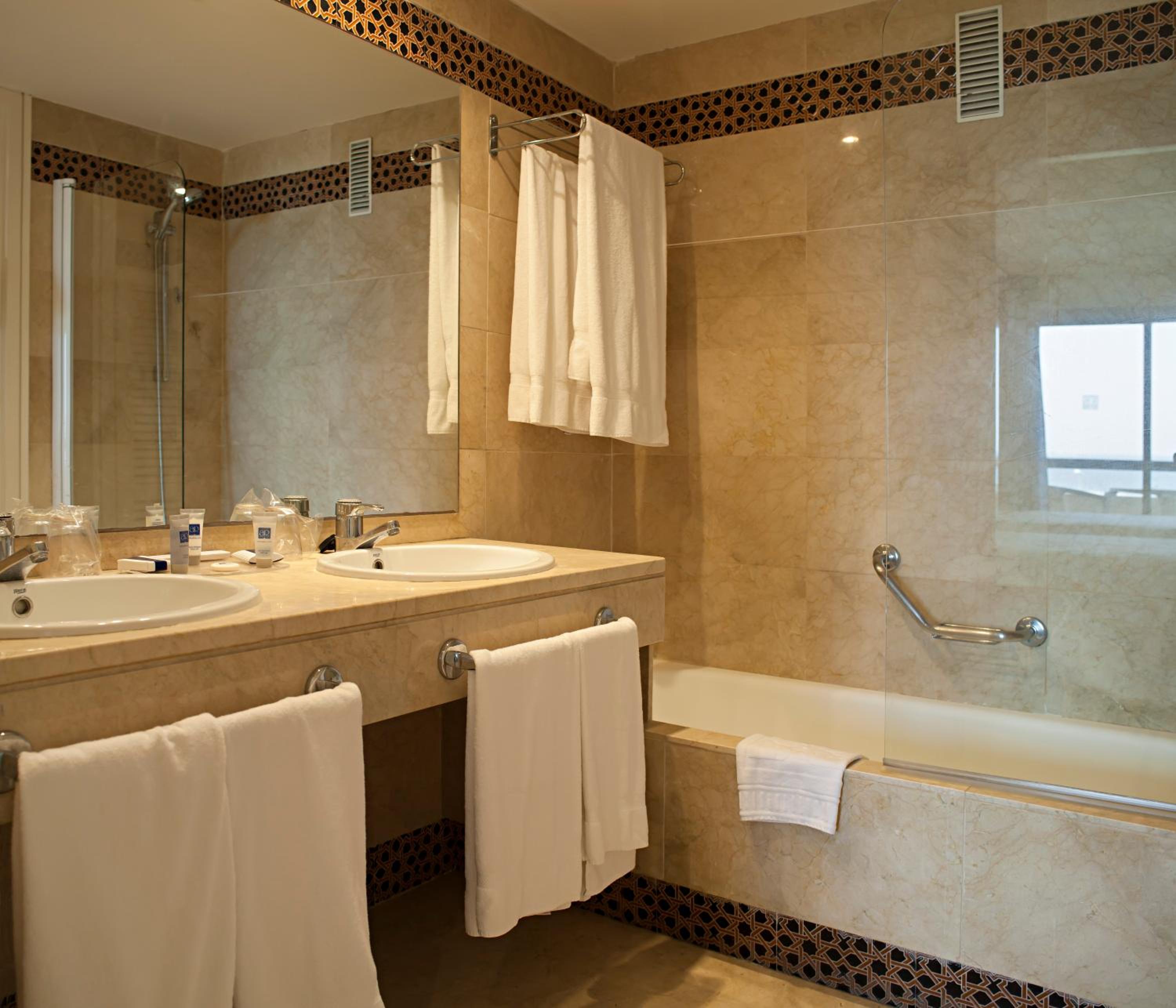 A marble-tiled bathroom with a long vanity, hanging white towels, and a bathtub with a glass screen.