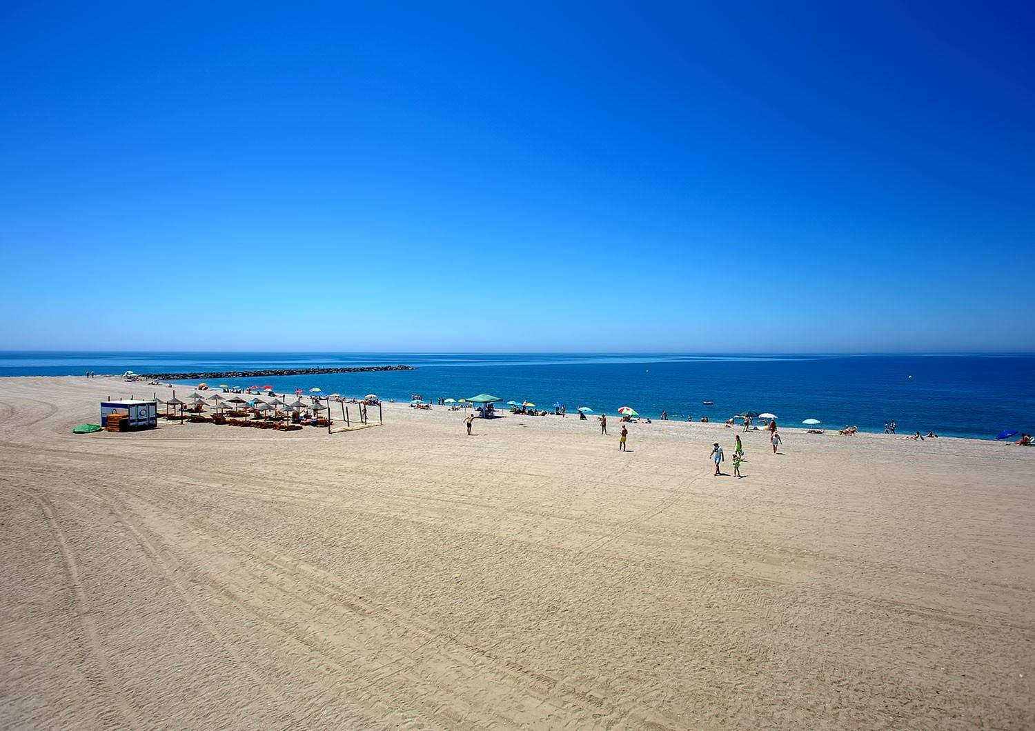A wide sandy beach under a clear blue sky, with sunshades and people along the calm sea.