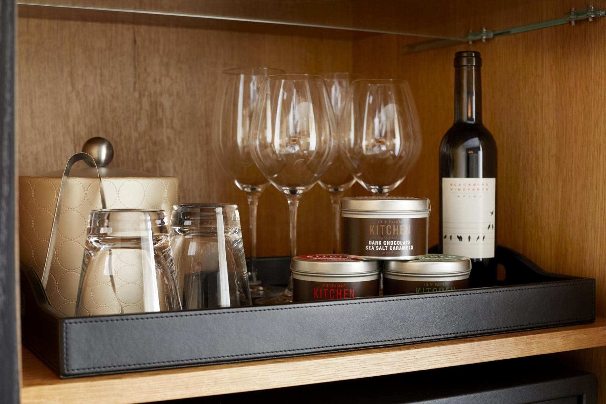 Wooden minibar shelf stocked with glassware, bottled wine, an ice bucket, and gourmet snack tins.