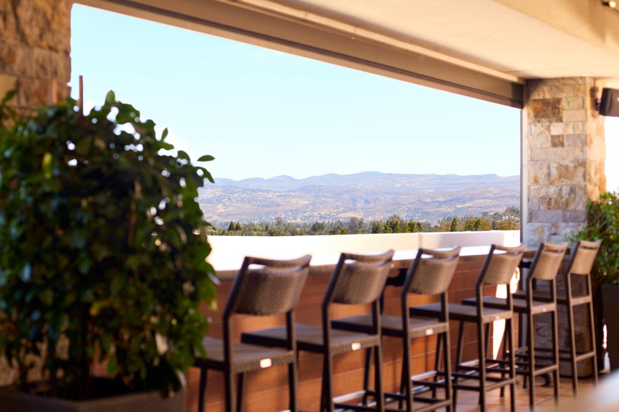 Open-air terrace bar with woven high chairs overlooking distant rolling hills and treetops under daylight.