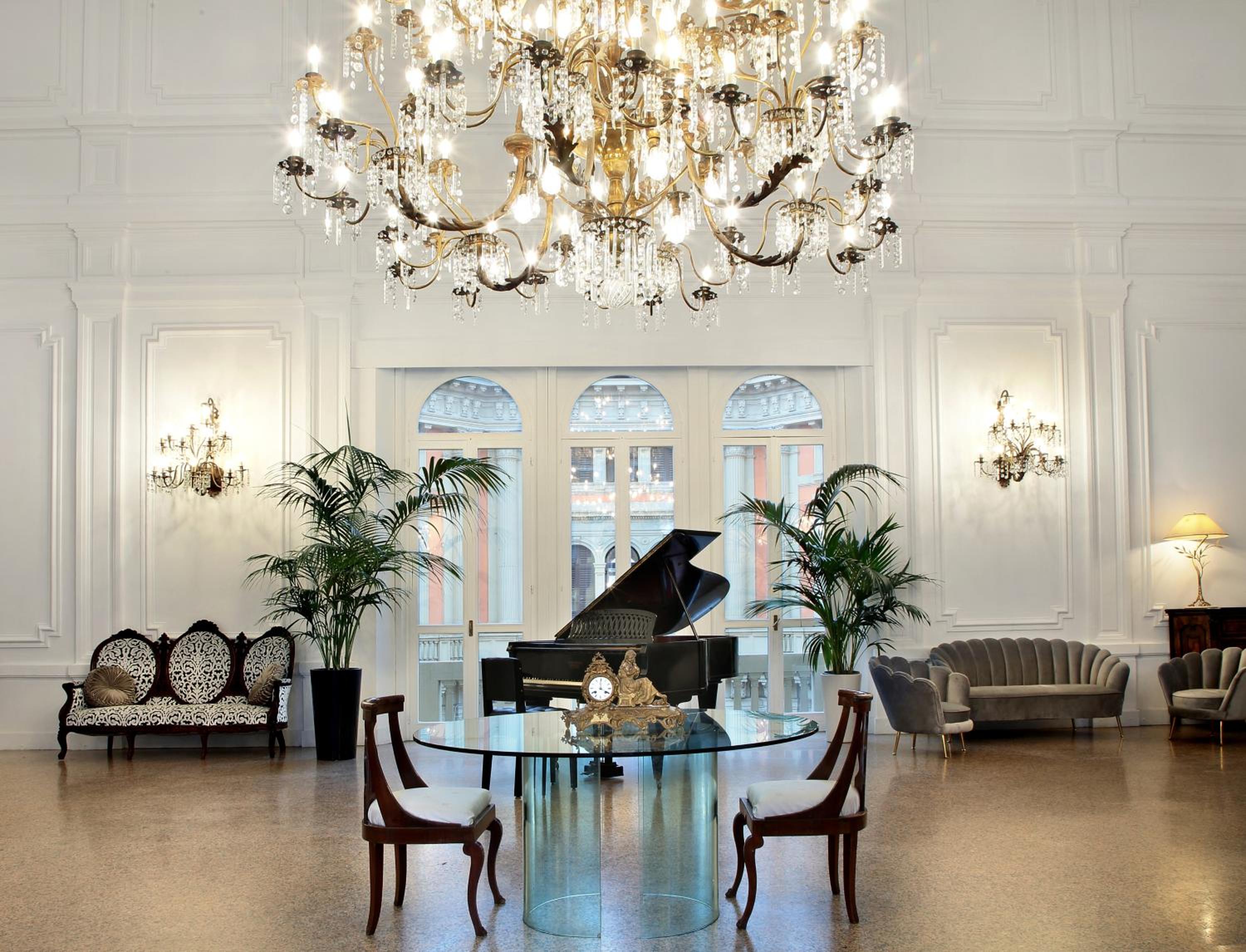 A white hall with high paneled walls, a large crystal chandelier, a grand piano, and potted palms.