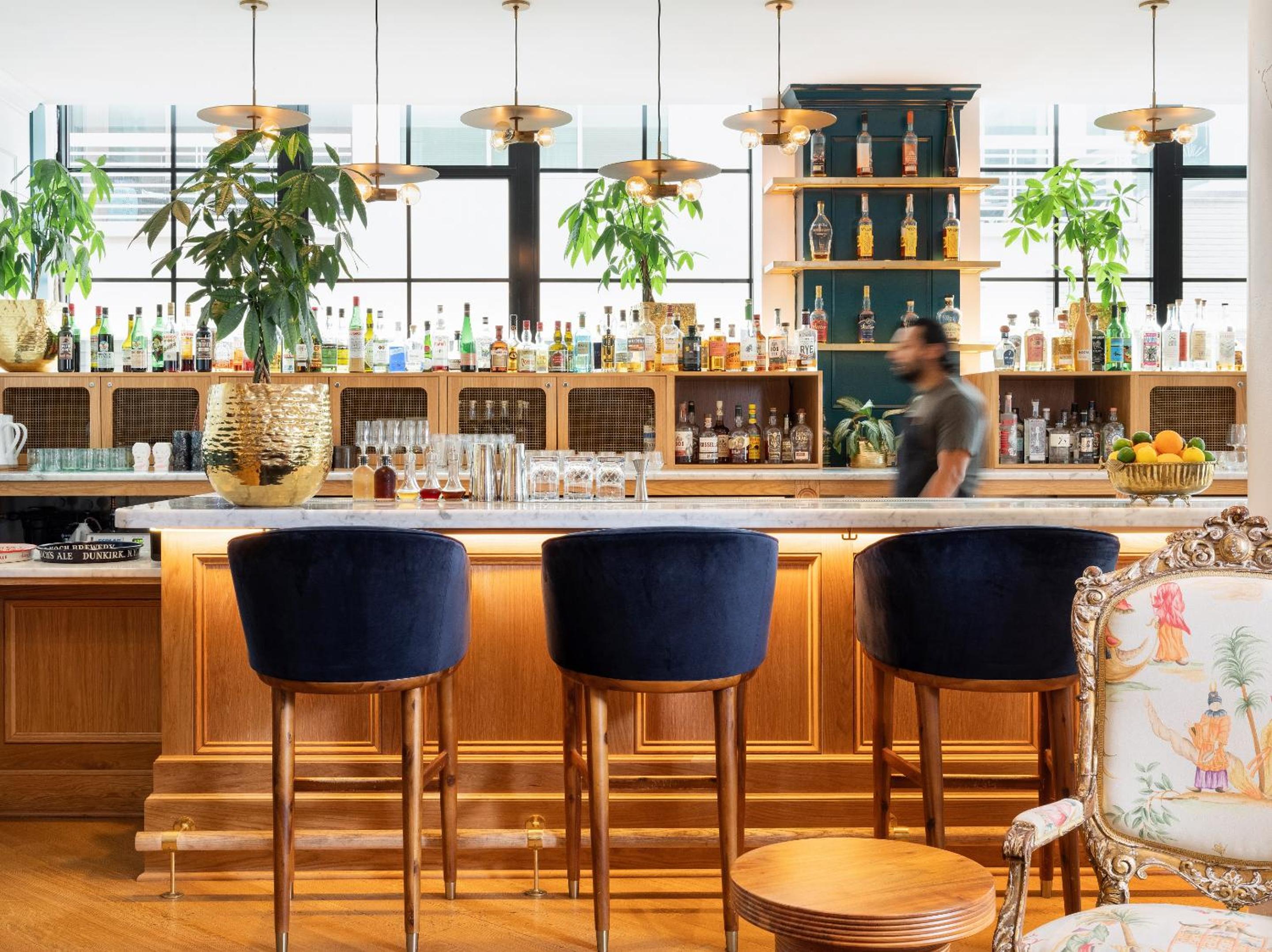 Blue velvet stools line a wooden bar with backlit liquor shelves and potted plants.
