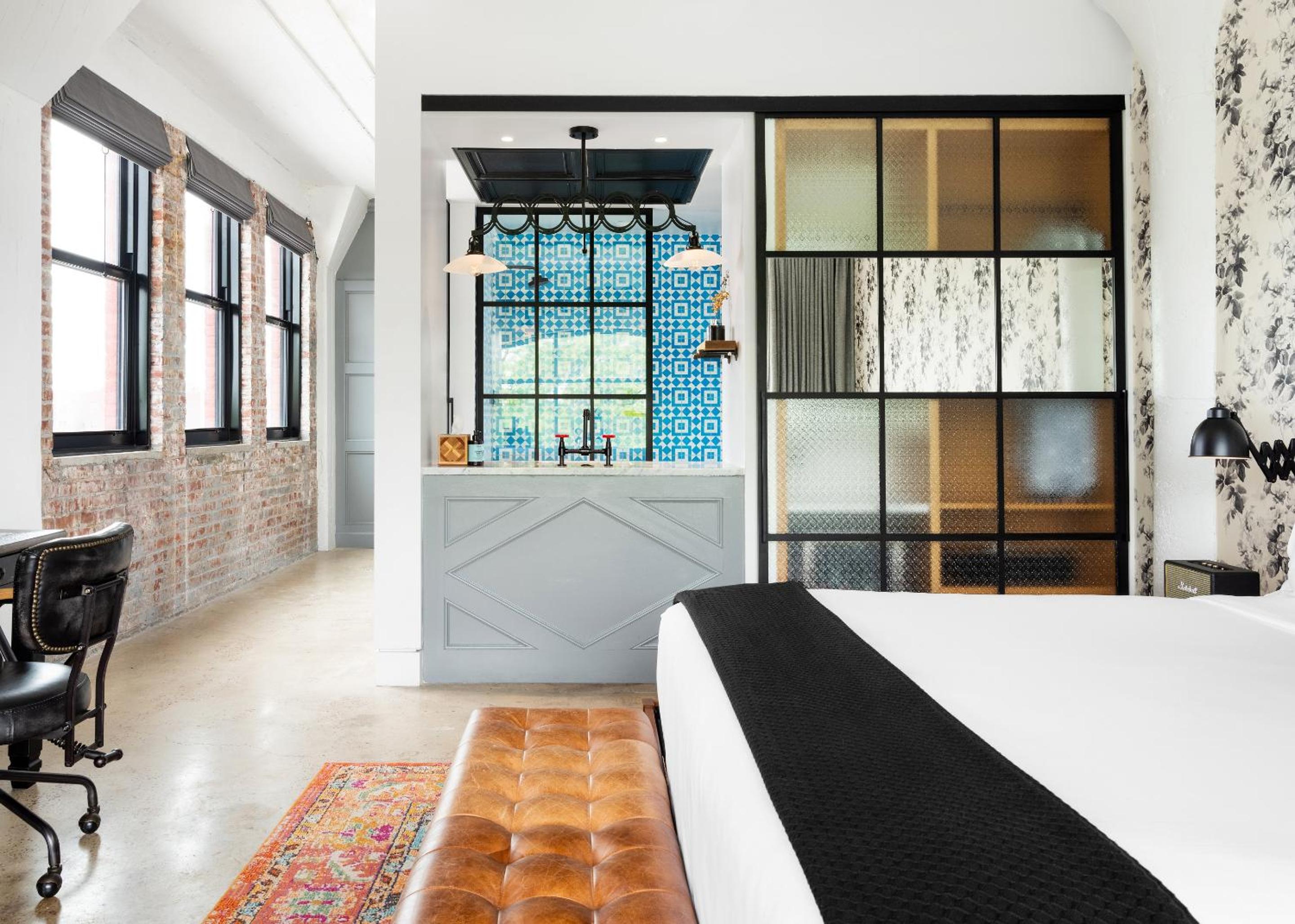 A bedroom featuring a leather bench at the foot of the bed, exposed brick wall, and glass-partitioned bathroom.