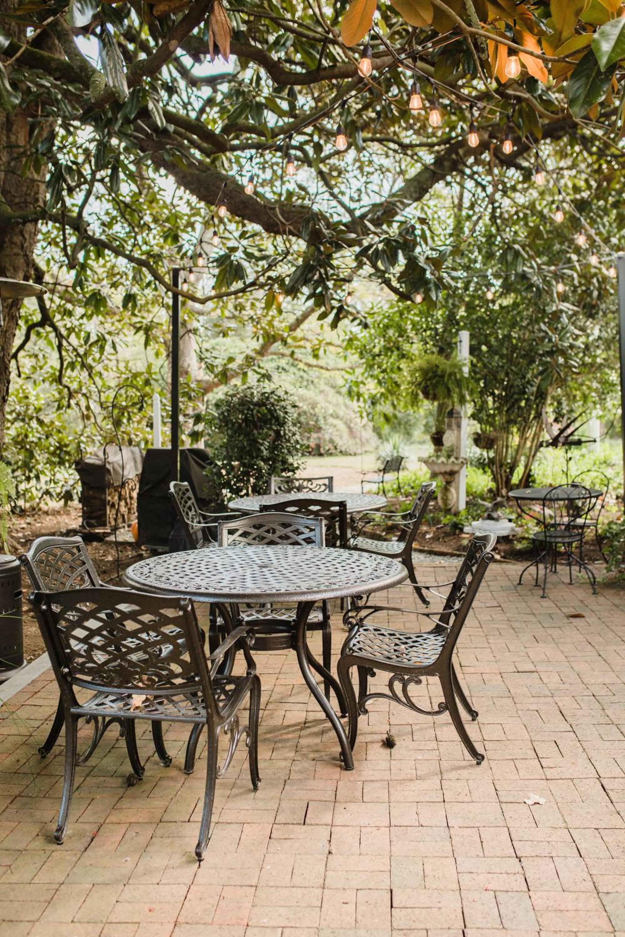 Dark wrought iron tables and chairs sit on a brick patio beneath trees draped with string lights.