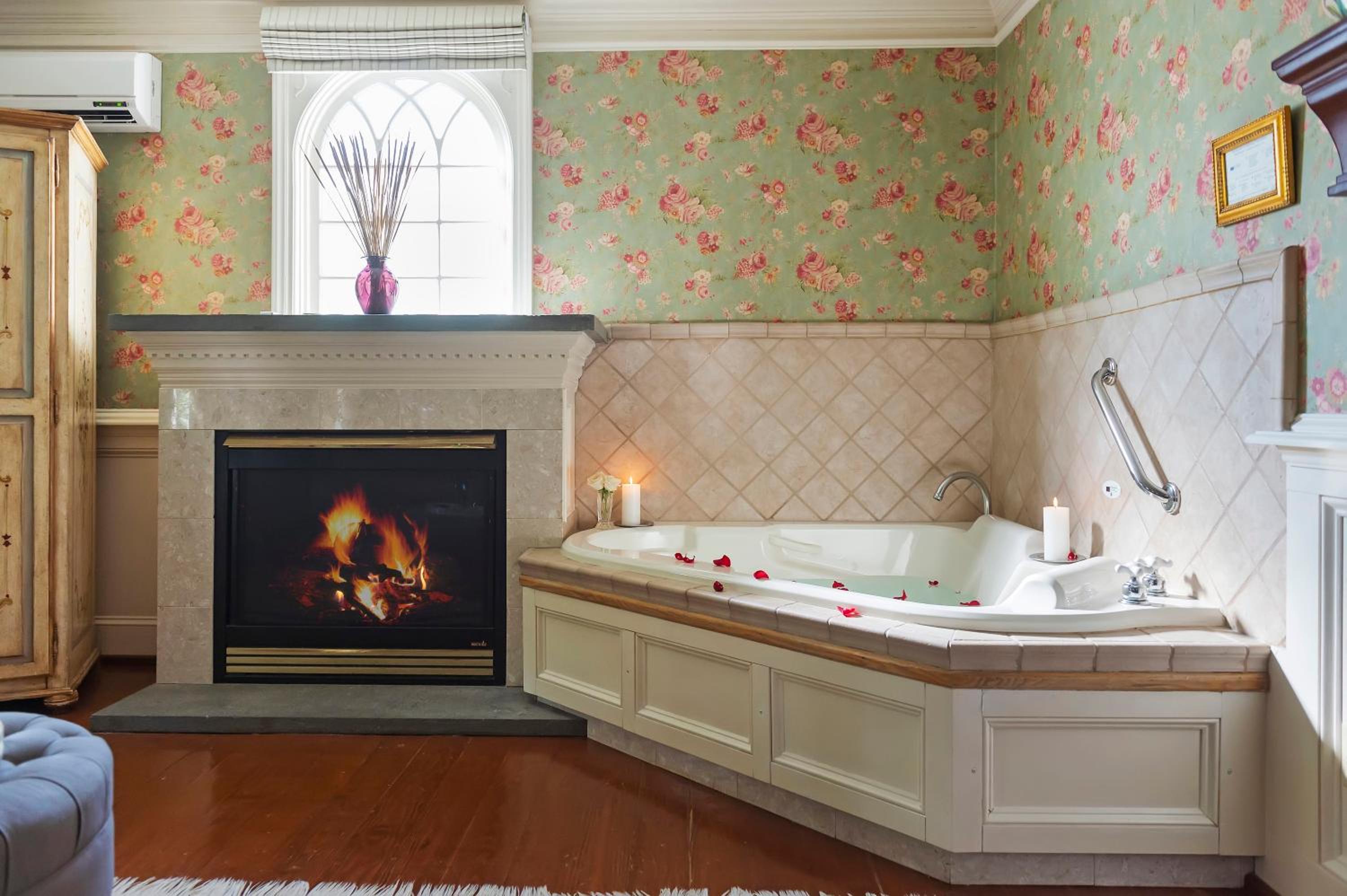 A corner jetted tub is set beside a fireplace with glowing flames beneath floral wallpaper.
