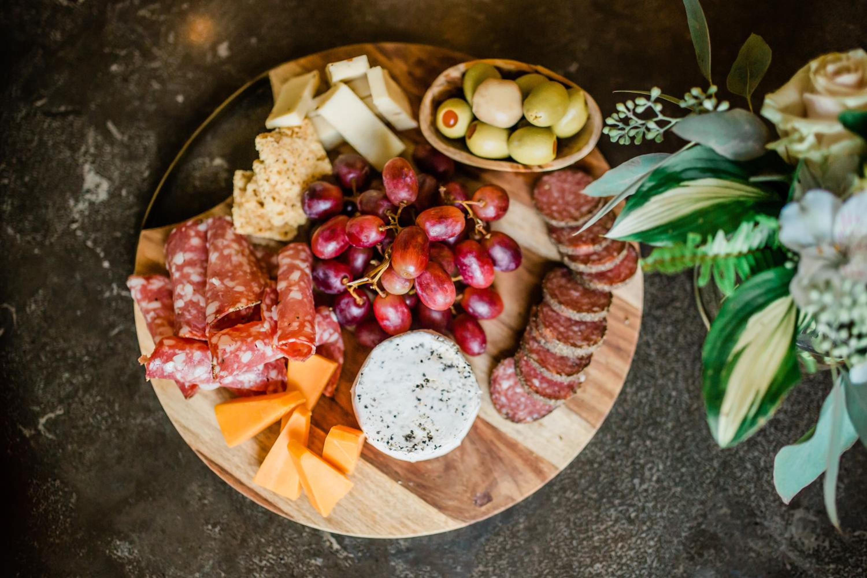 A round wooden board displays cured meats, cheese wedges, dark grapes, and green olives beside fresh flowers.