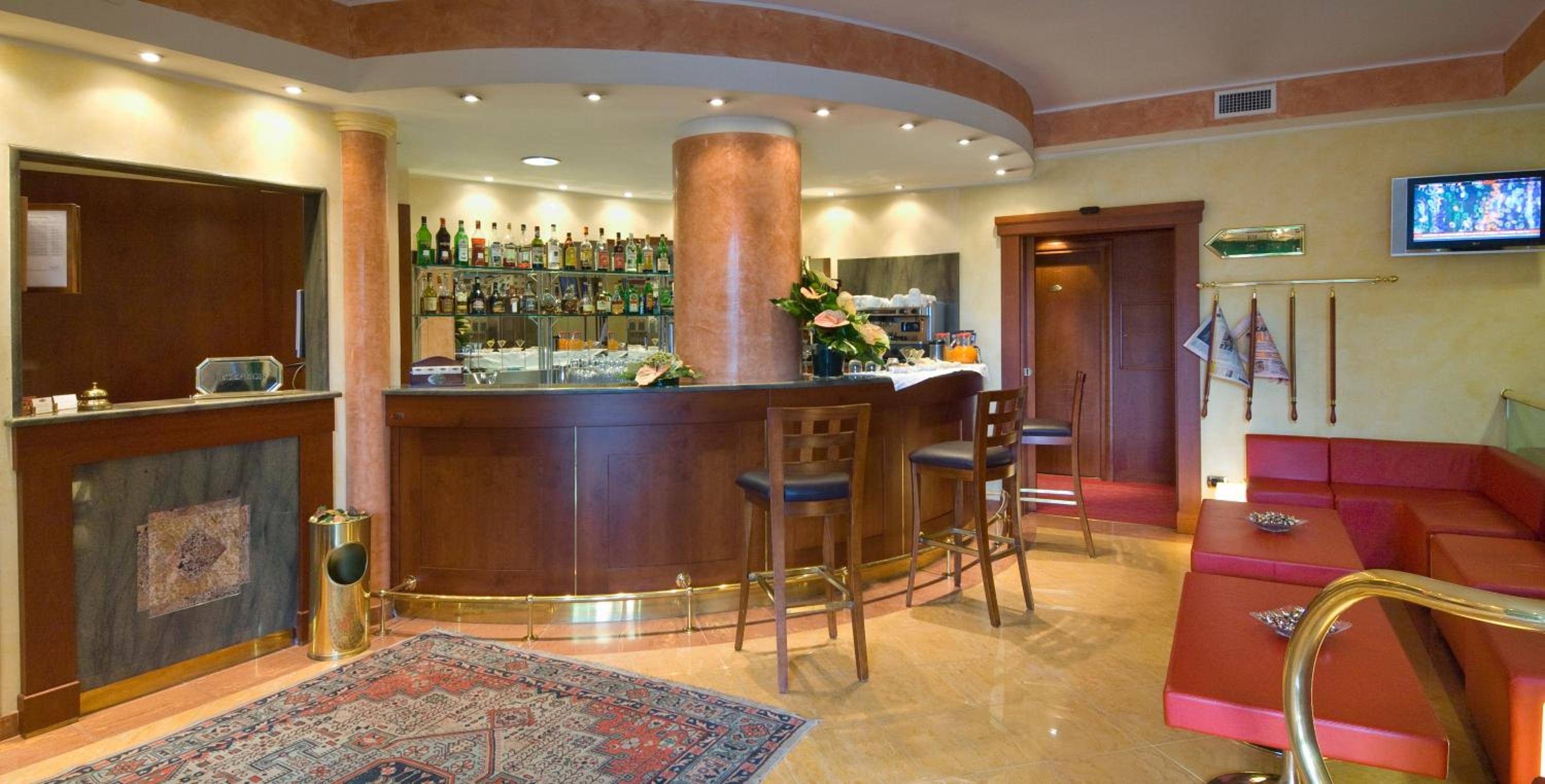 A hotel bar features a curved wooden counter, tall barstools, an ornate rug, and red bench seating.