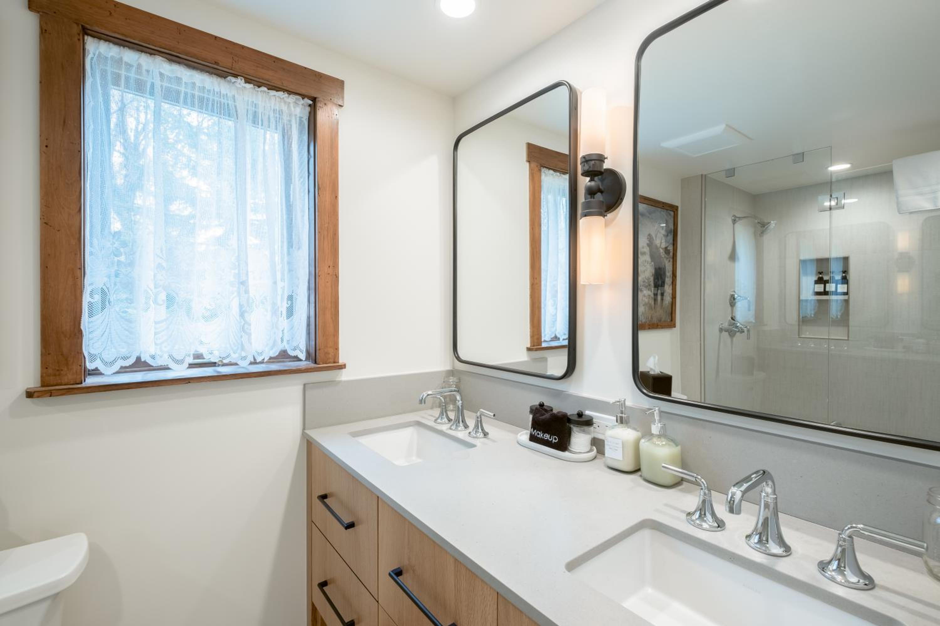 Modern bathroom vanity with stone countertop, black-framed mirrors, and a window framed in wood with lace curtains.