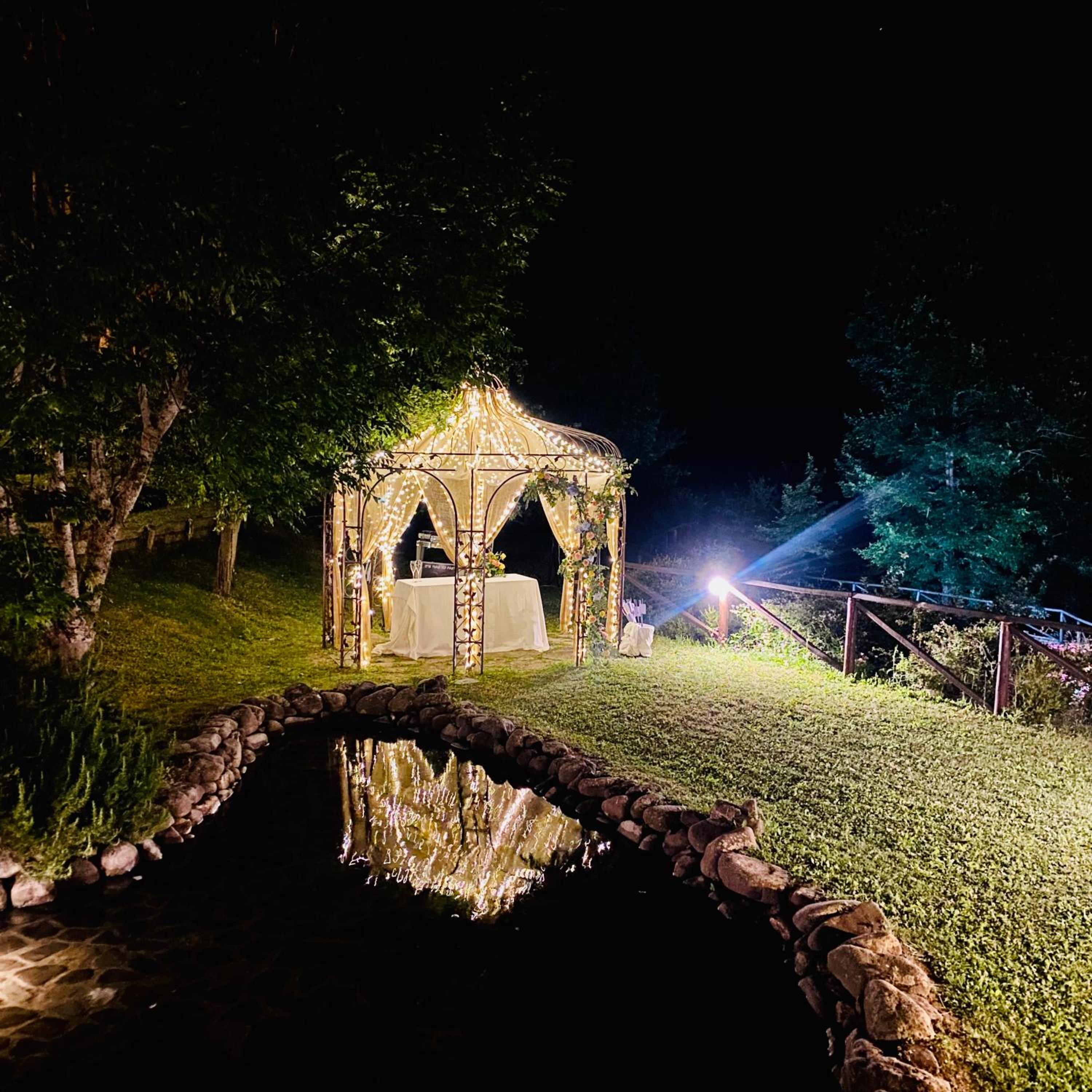 An illuminated gazebo decorated with fairy lights reflects in a stone-lined pond at night.