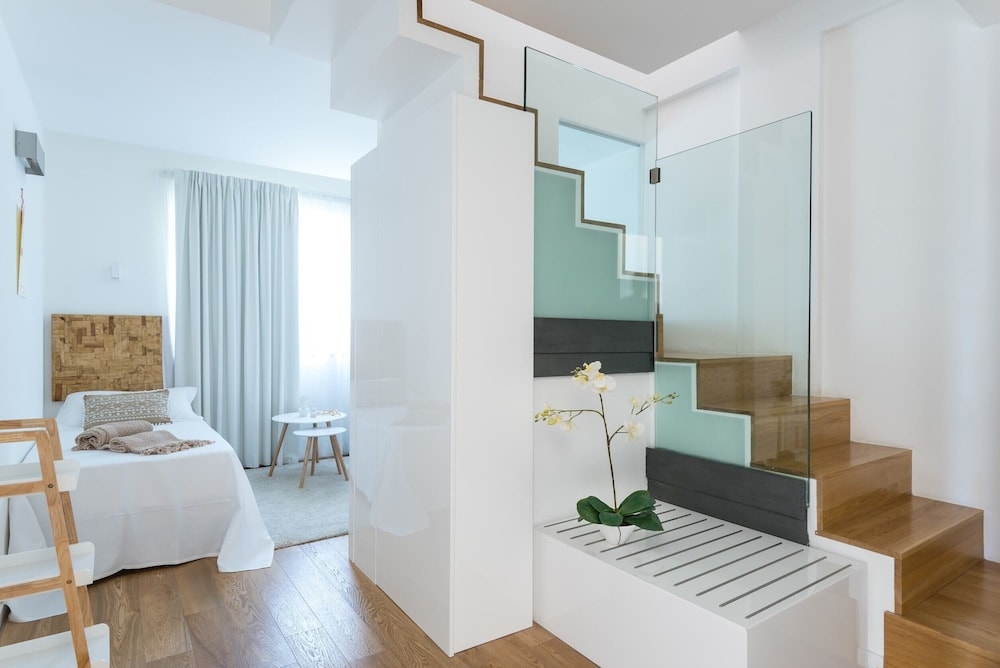 White bedroom with wood floors, featuring a bed, side table, and a wooden staircase with glass railings.