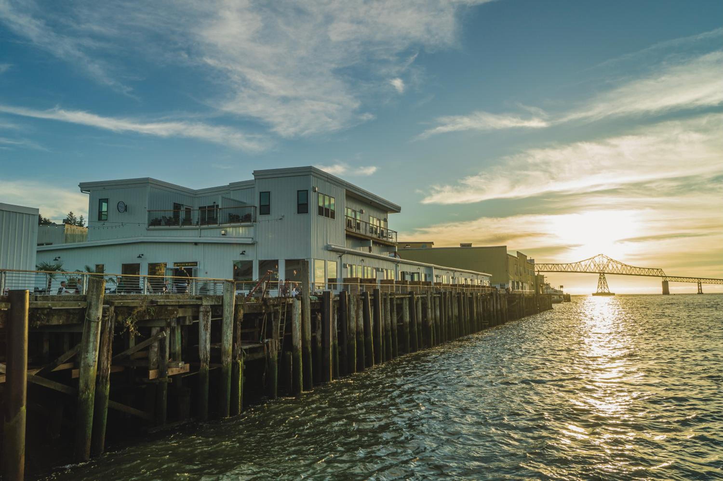 Waterfront hotel built on wooden pilings over the river at sunset, with a truss bridge in the distance.