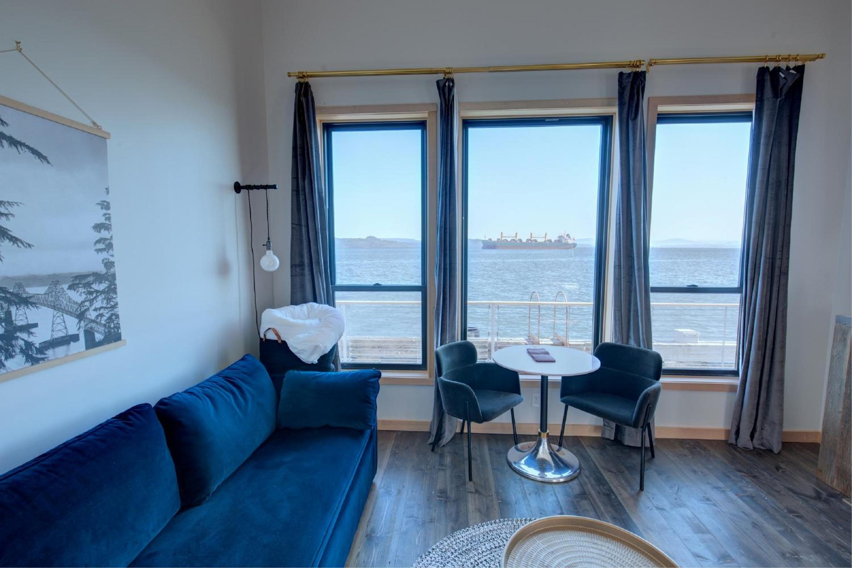 A seating area with a blue velvet sofa and armchairs fronting windows overlooking river water and a cargo ship.