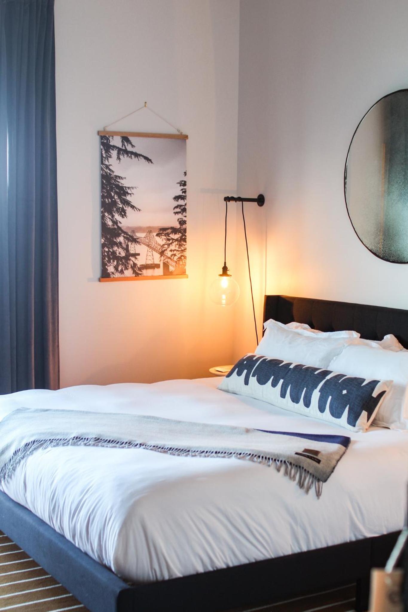 A hotel bed made with white linens and a patterned bolster pillow, illuminated by a warm bedside pendant light.