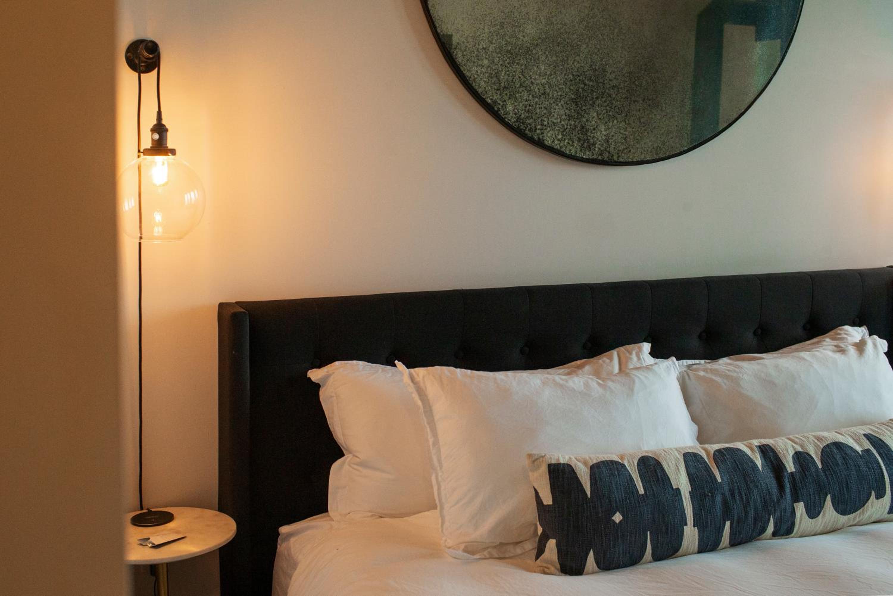 A dark tufted headboard behind white pillows and a patterned bolster, beside a glowing glass pendant lamp.
