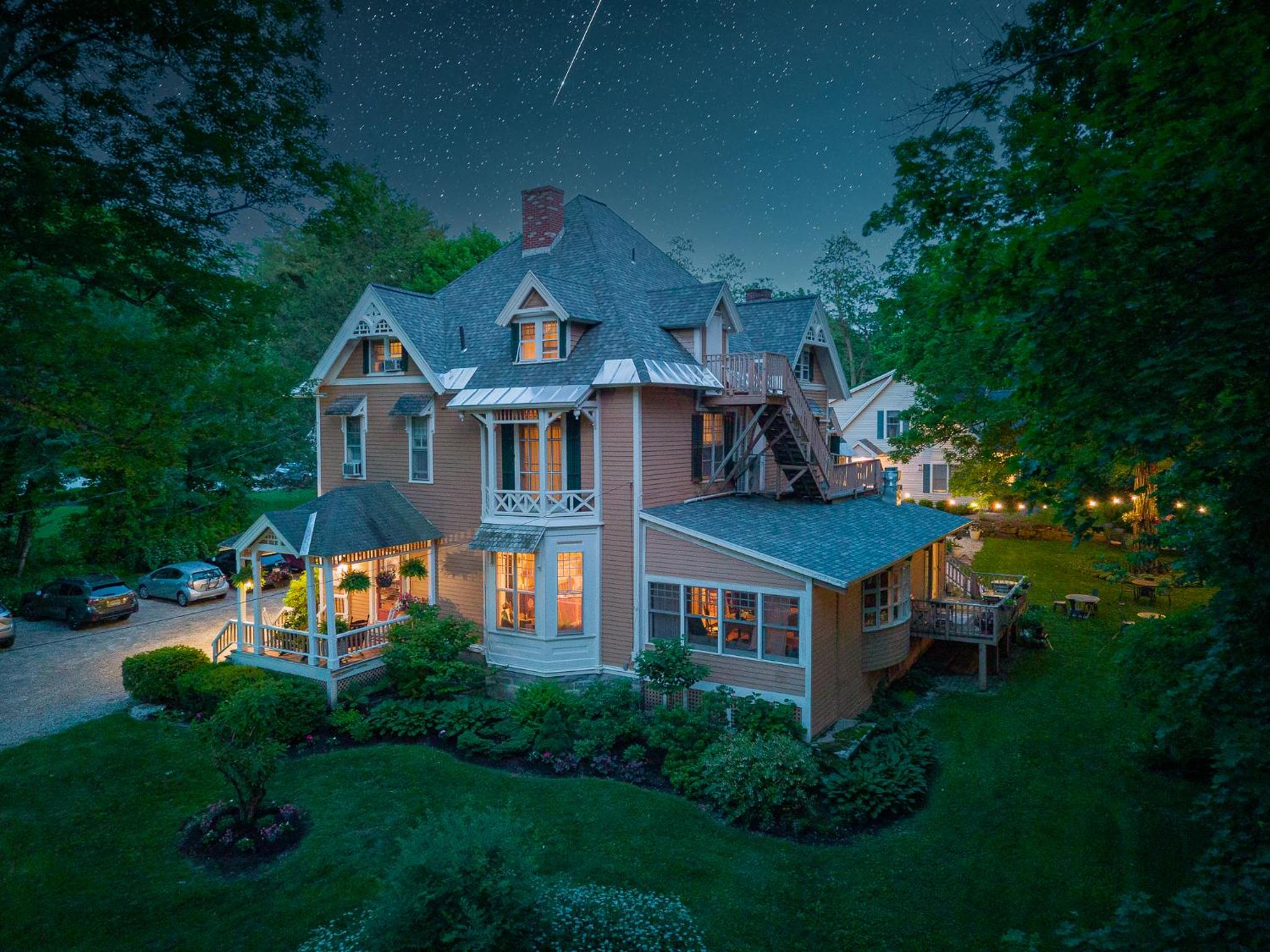 Exterior night view of the Victorian inn lit from within, surrounded by lawn and trees under a starry sky.