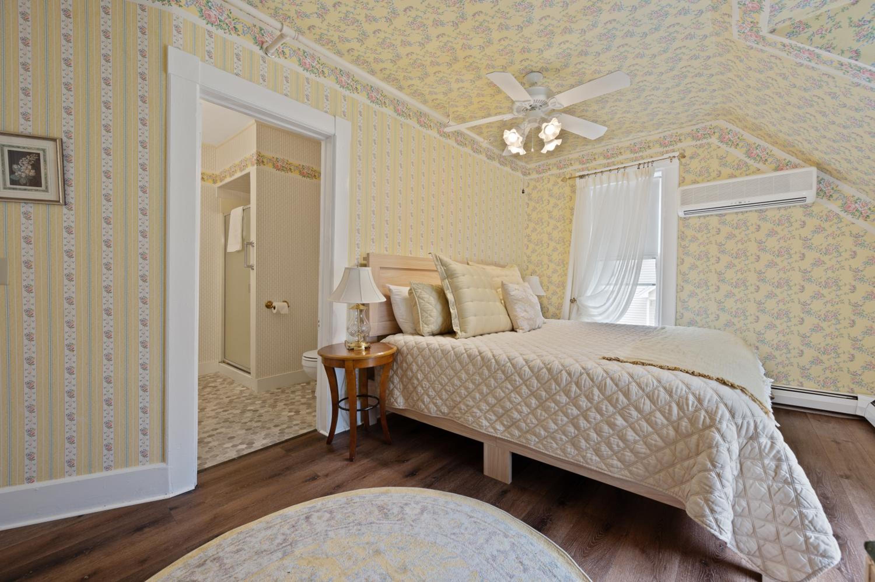 A bedroom with yellow floral wallpaper, sloping ceiling, wood floor, and doorway opening into a bathroom.