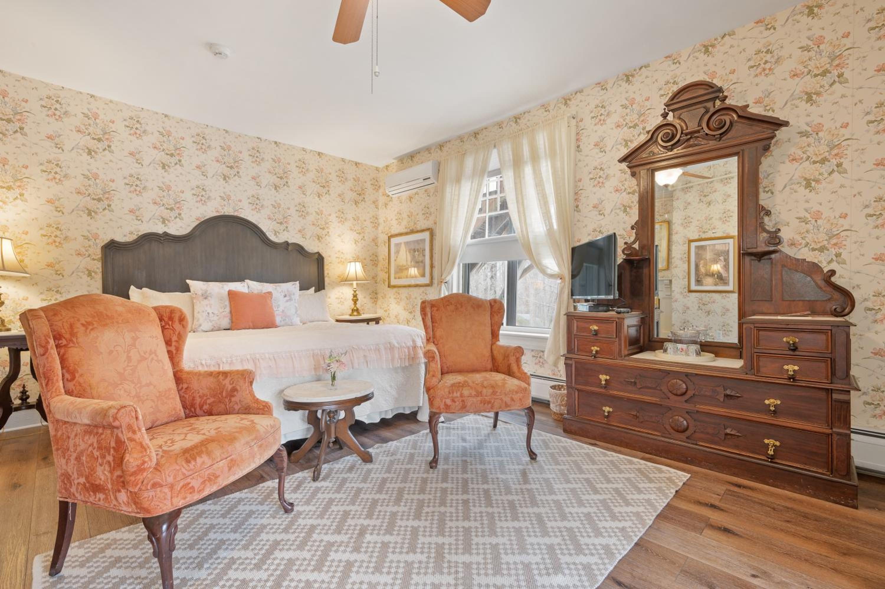 A guest room with floral wallpaper, an ornate carved wooden dresser and mirror, upholstered armchairs, and a bed.