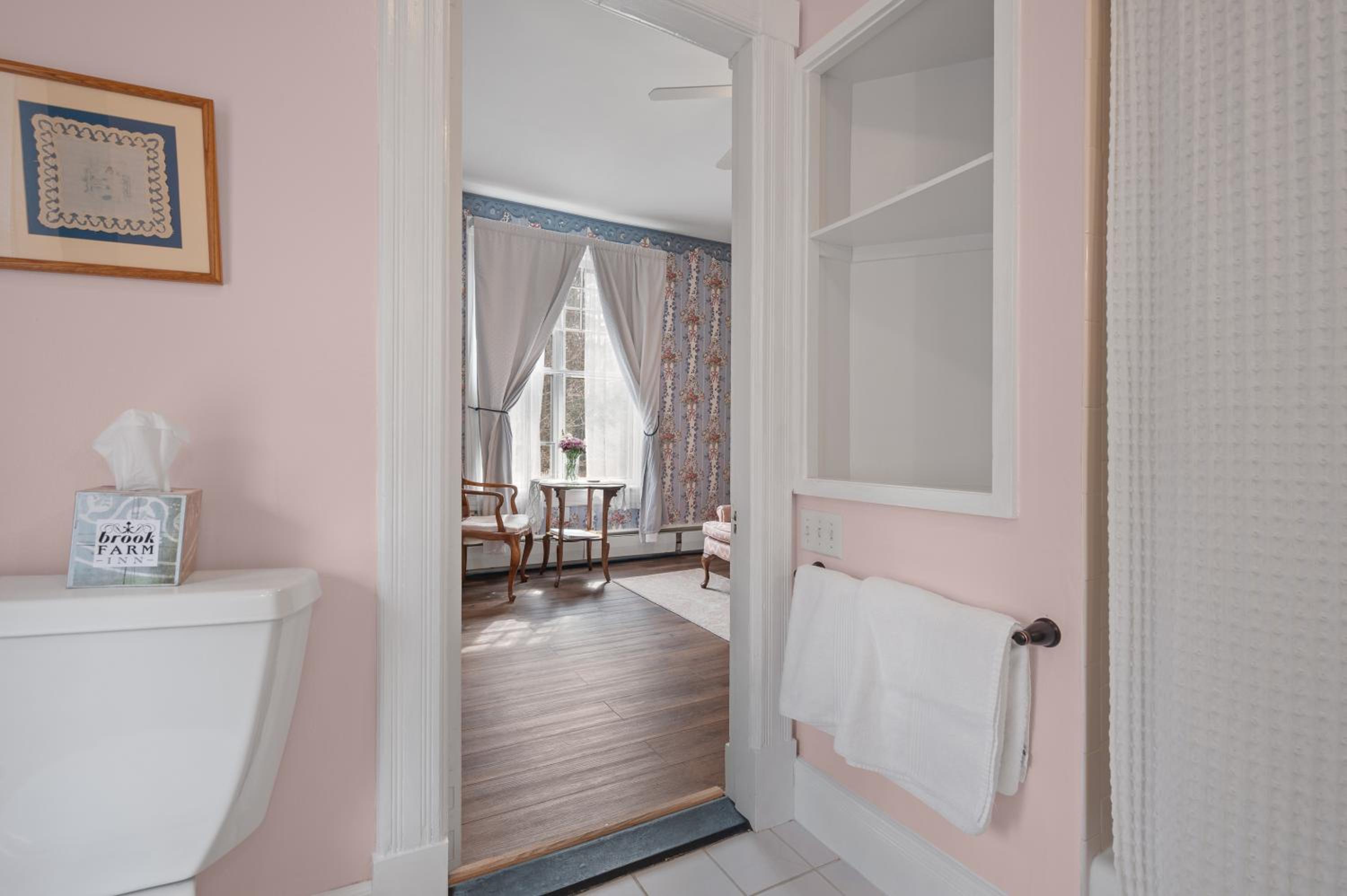 View looking from a soft pink bathroom with folded white towels into a bedroom with tall draped windows.