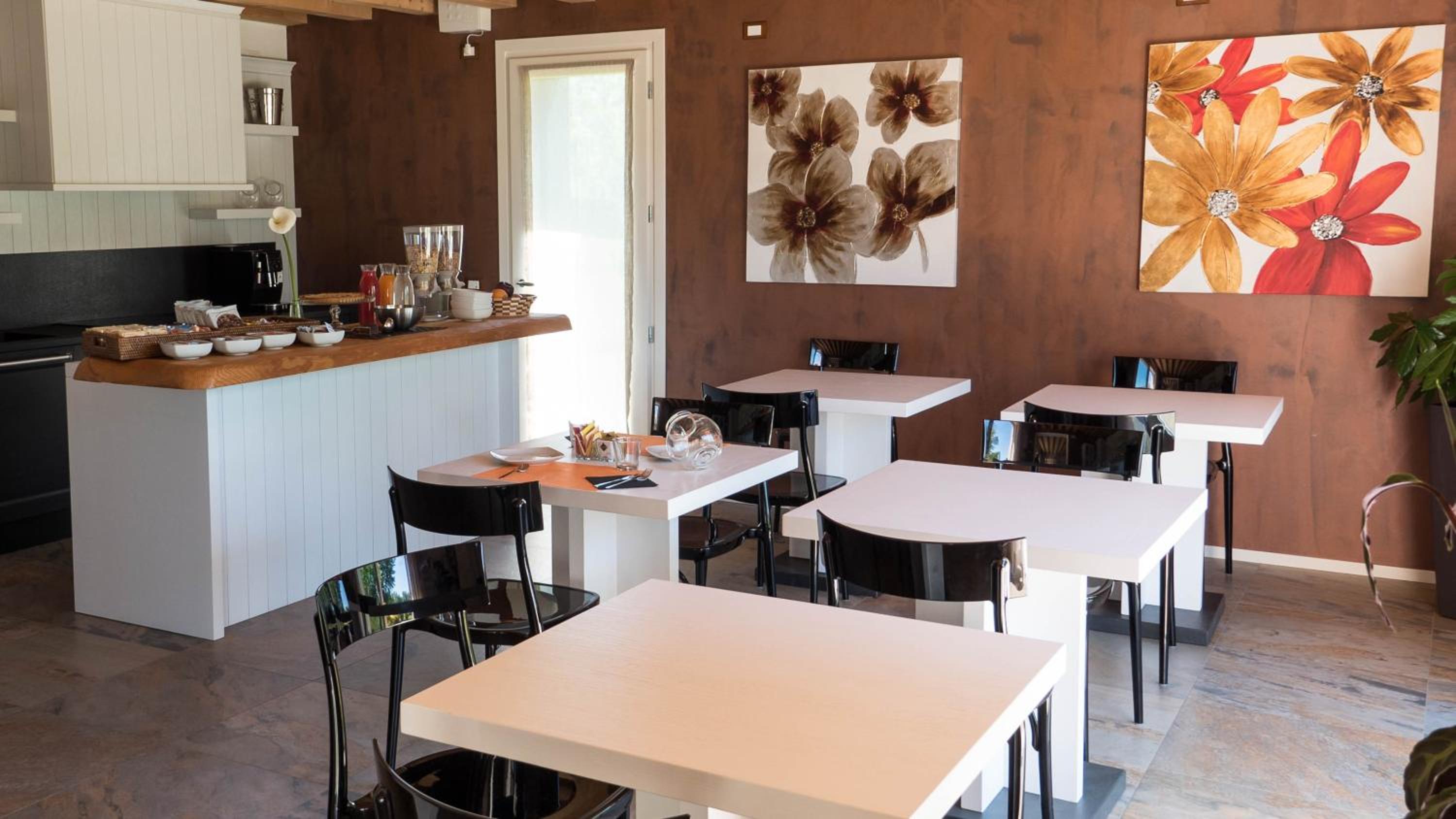 An indoor dining area with white tables, glossy black chairs, floral artwork, and a breakfast buffet counter.