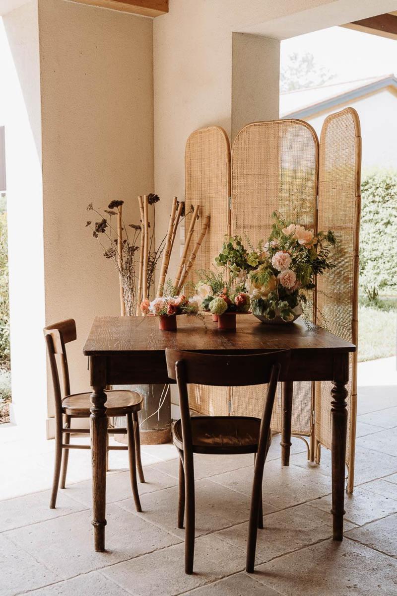 A wooden table with chairs and floral arrangements set before a woven screen under a covered patio.