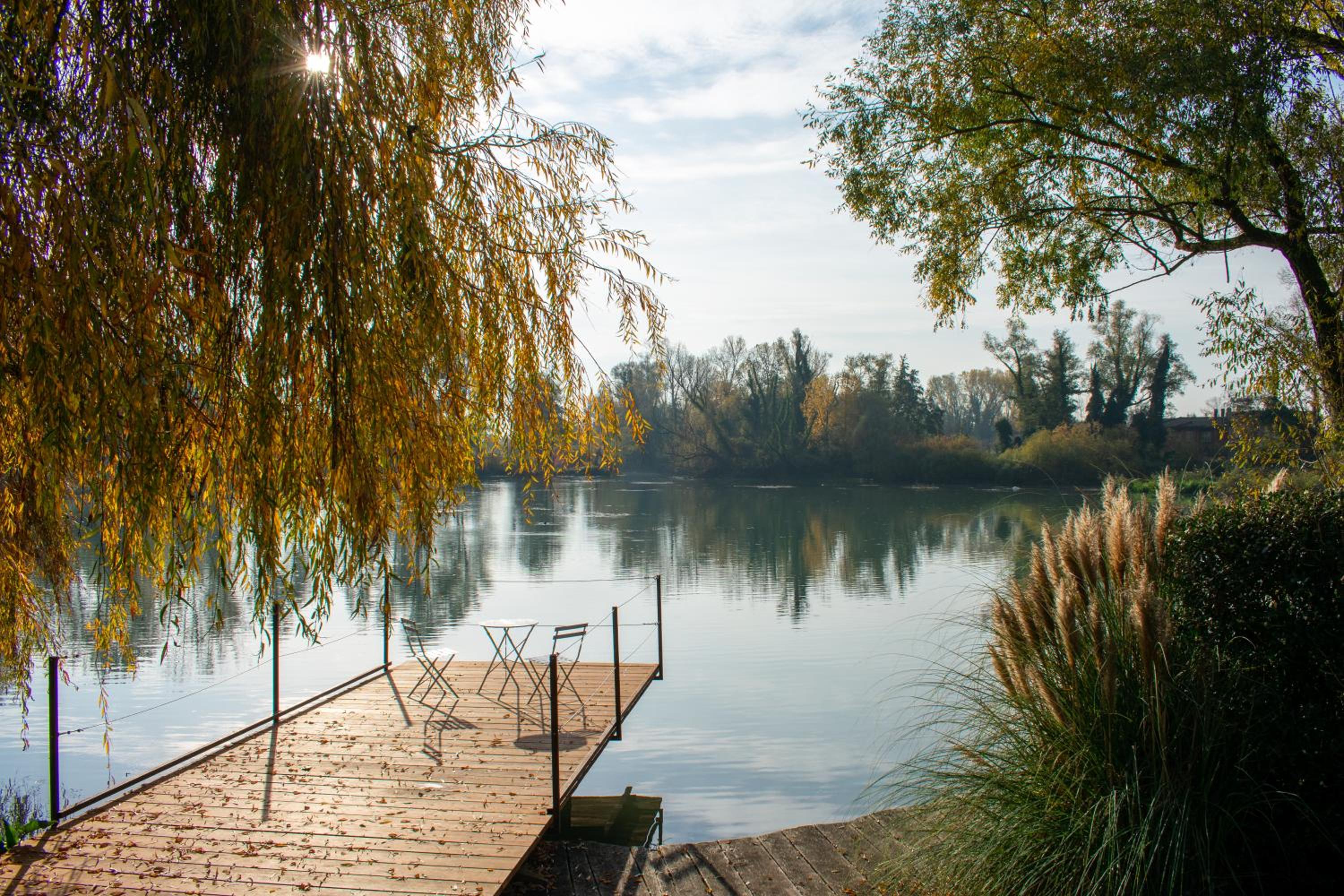 A wooden jetty with a small table and chairs extending into a calm river under weeping willow branches.