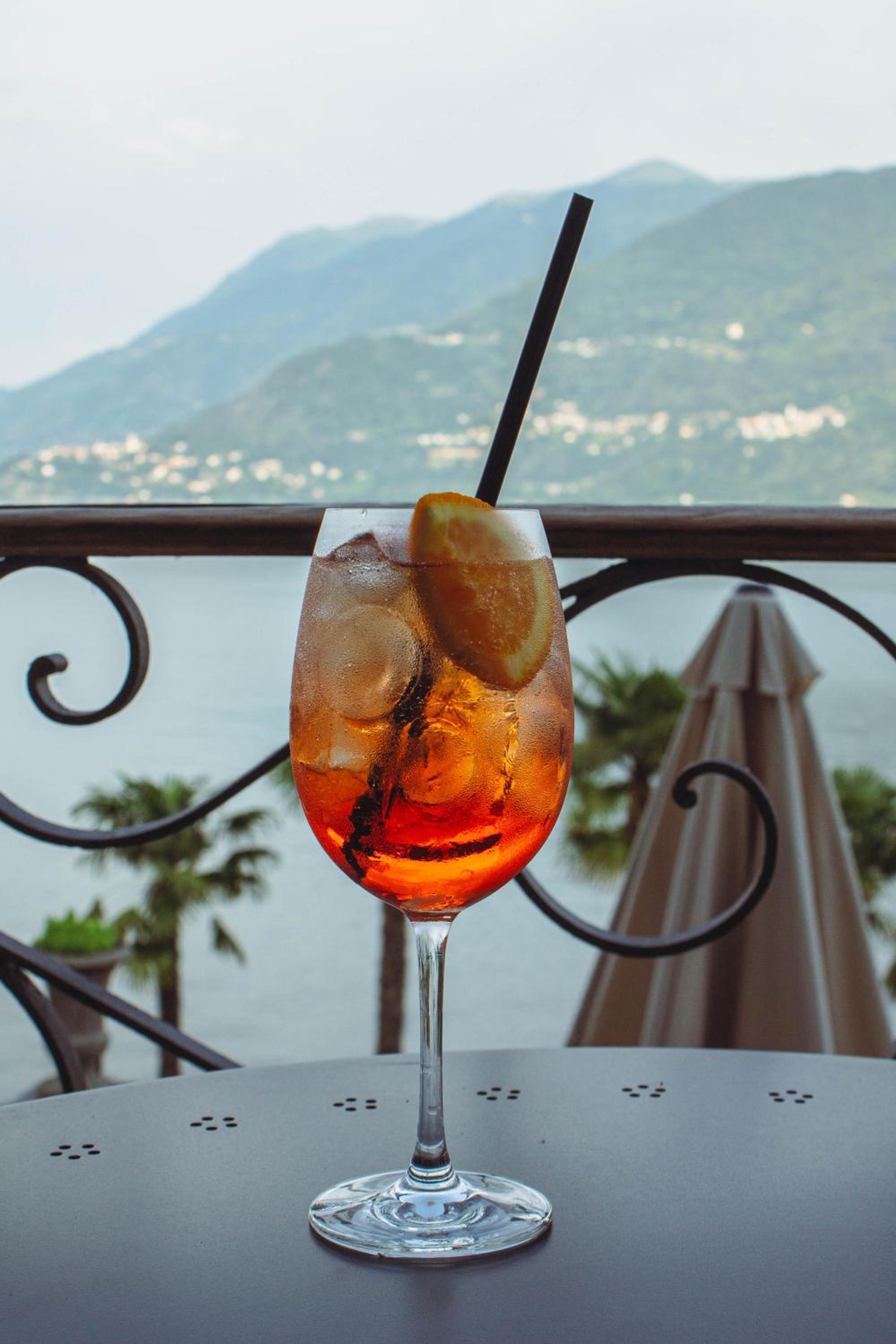 An iced spritz garnished with an orange slice sits on a terrace table overlooking a lake and distant mountains.