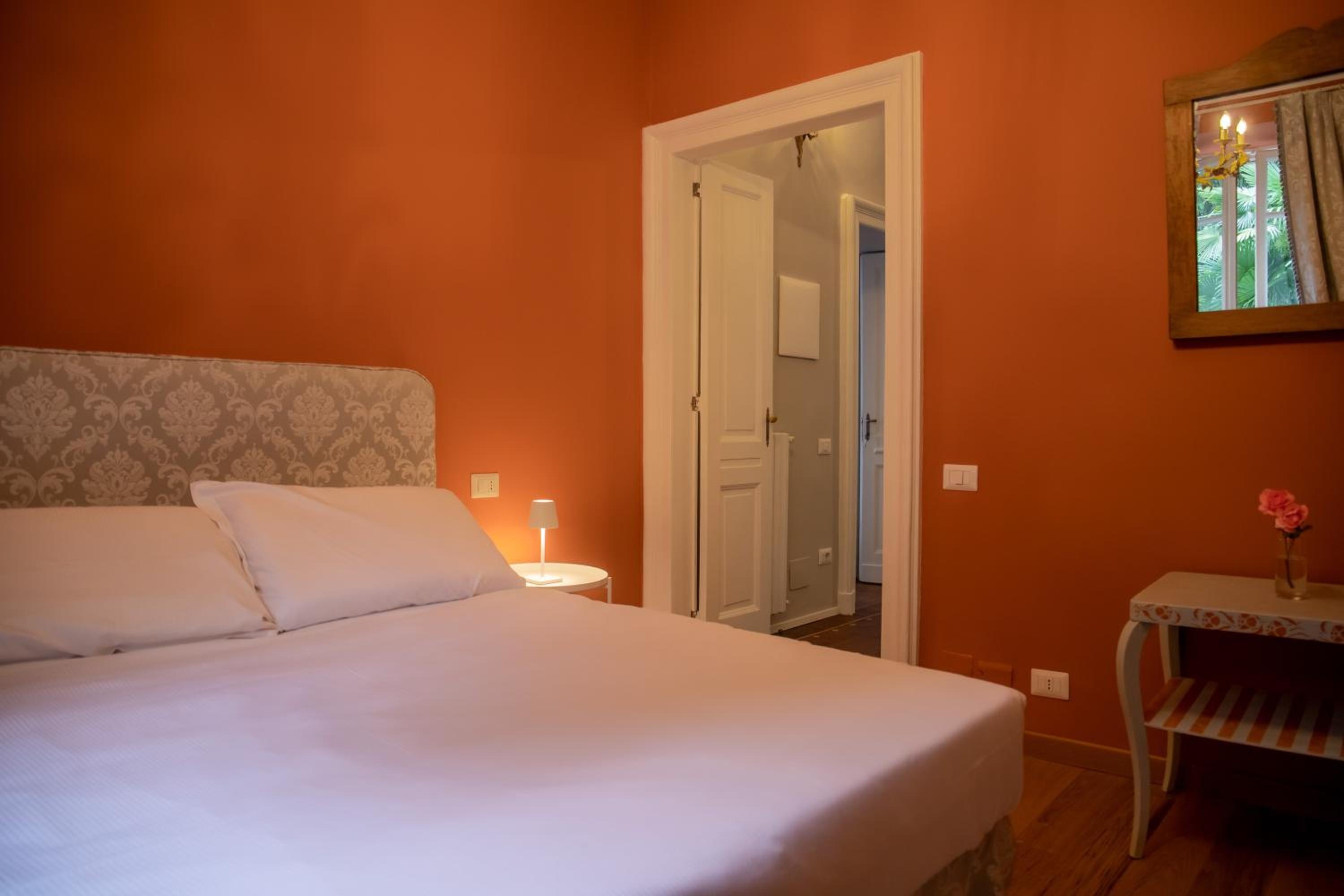A bedroom with terracotta orange walls, a double bed with a brocade headboard, and an open doorway to a hallway.