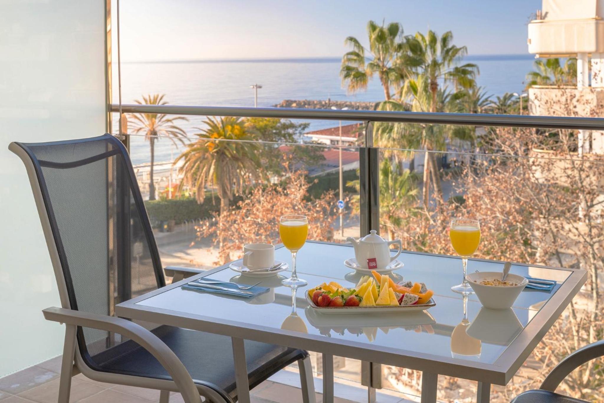 Juice, sliced fruit, and tea served on a glass balcony table overlooking palm trees and the sea.