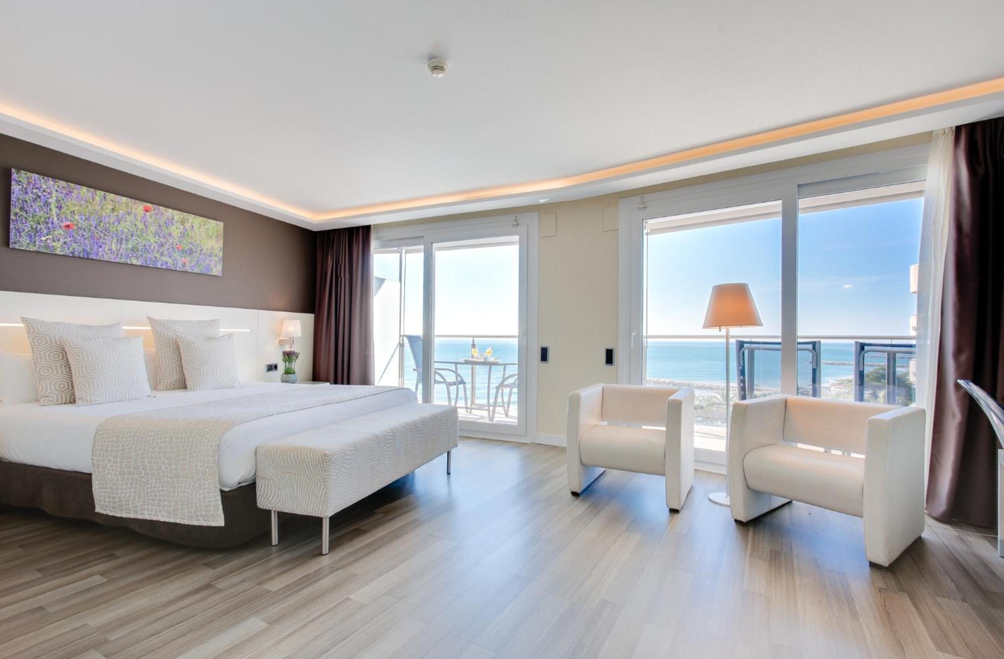 Bedroom with wood-look flooring, white armchairs, and sliding glass doors opening to a sea view.