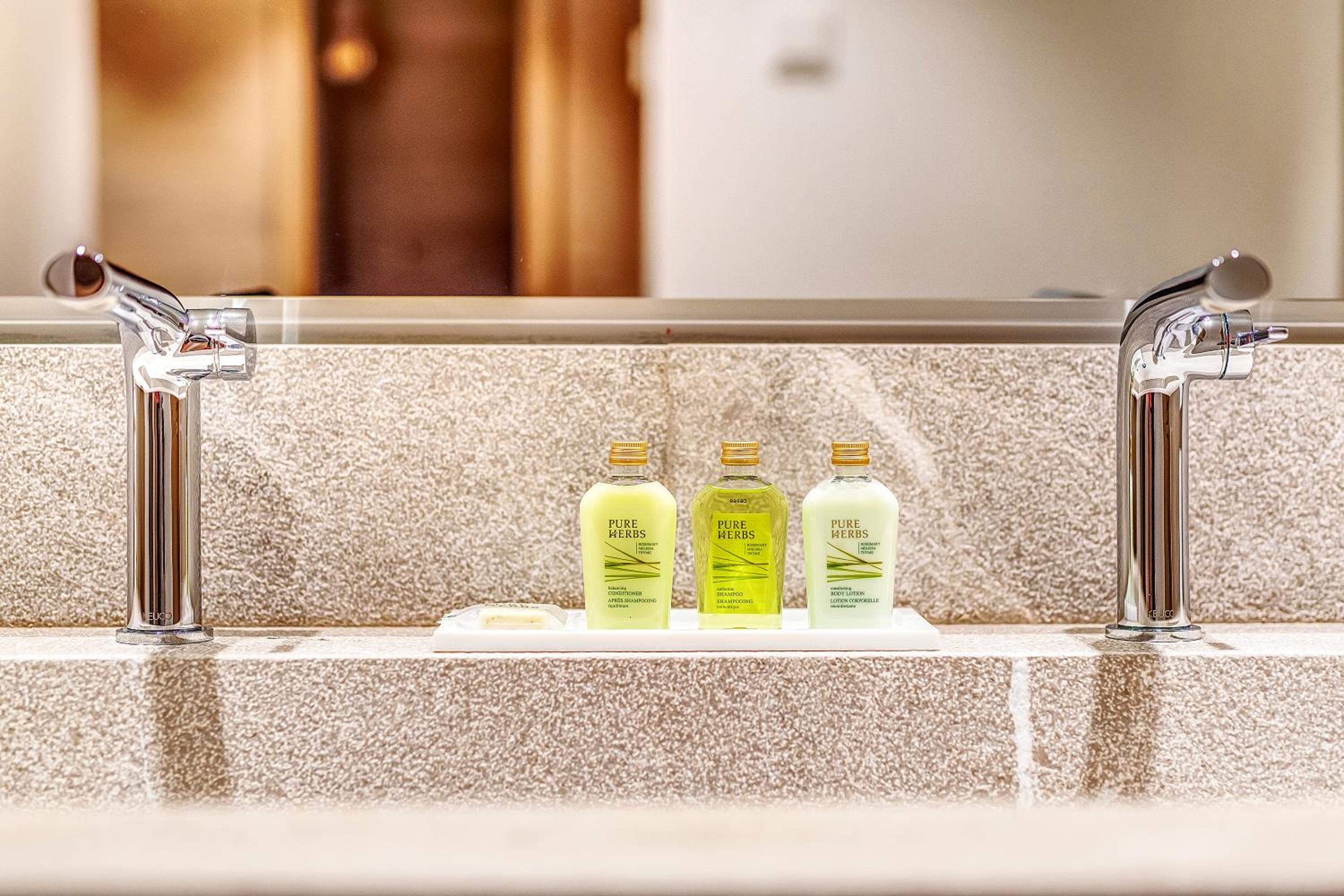Toiletries tray set between chrome faucets on a stone countertop in front of a mirror.