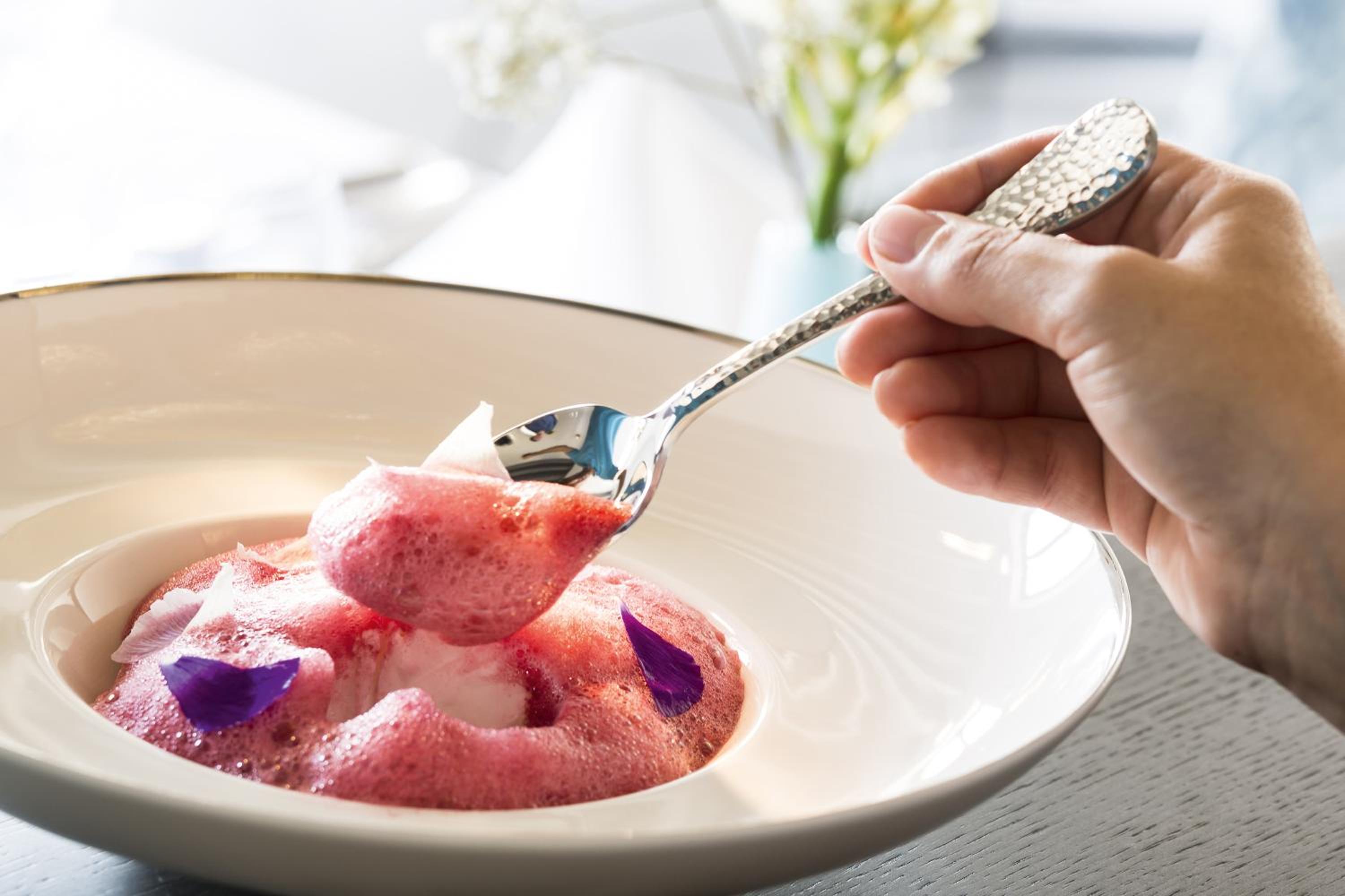 A spoon lifting pink dessert foam from a wide white bowl garnished with purple flower petals.