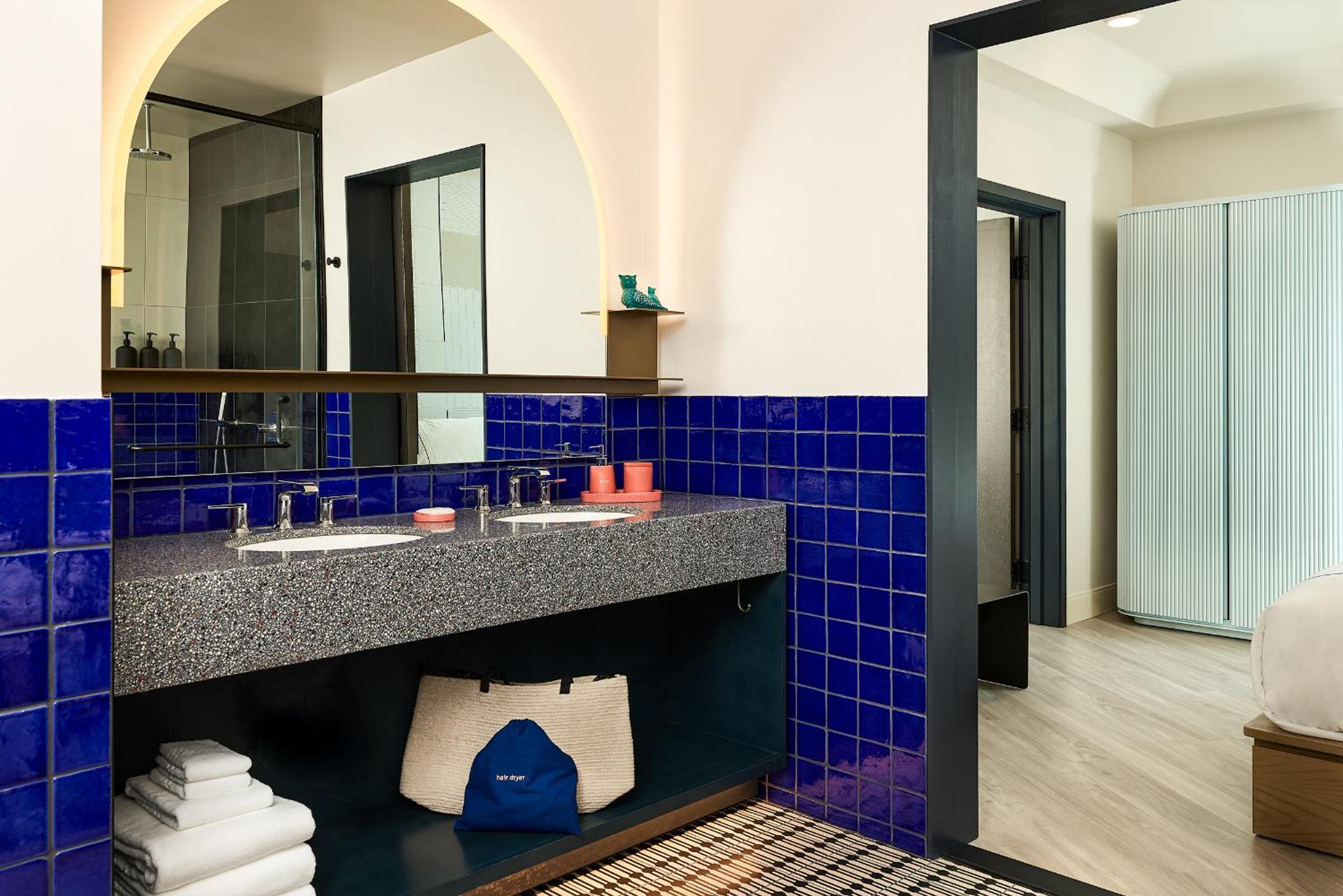 A bathroom with cobalt blue wall tiles, a speckled stone countertop, an arched mirror, and patterned tile flooring.