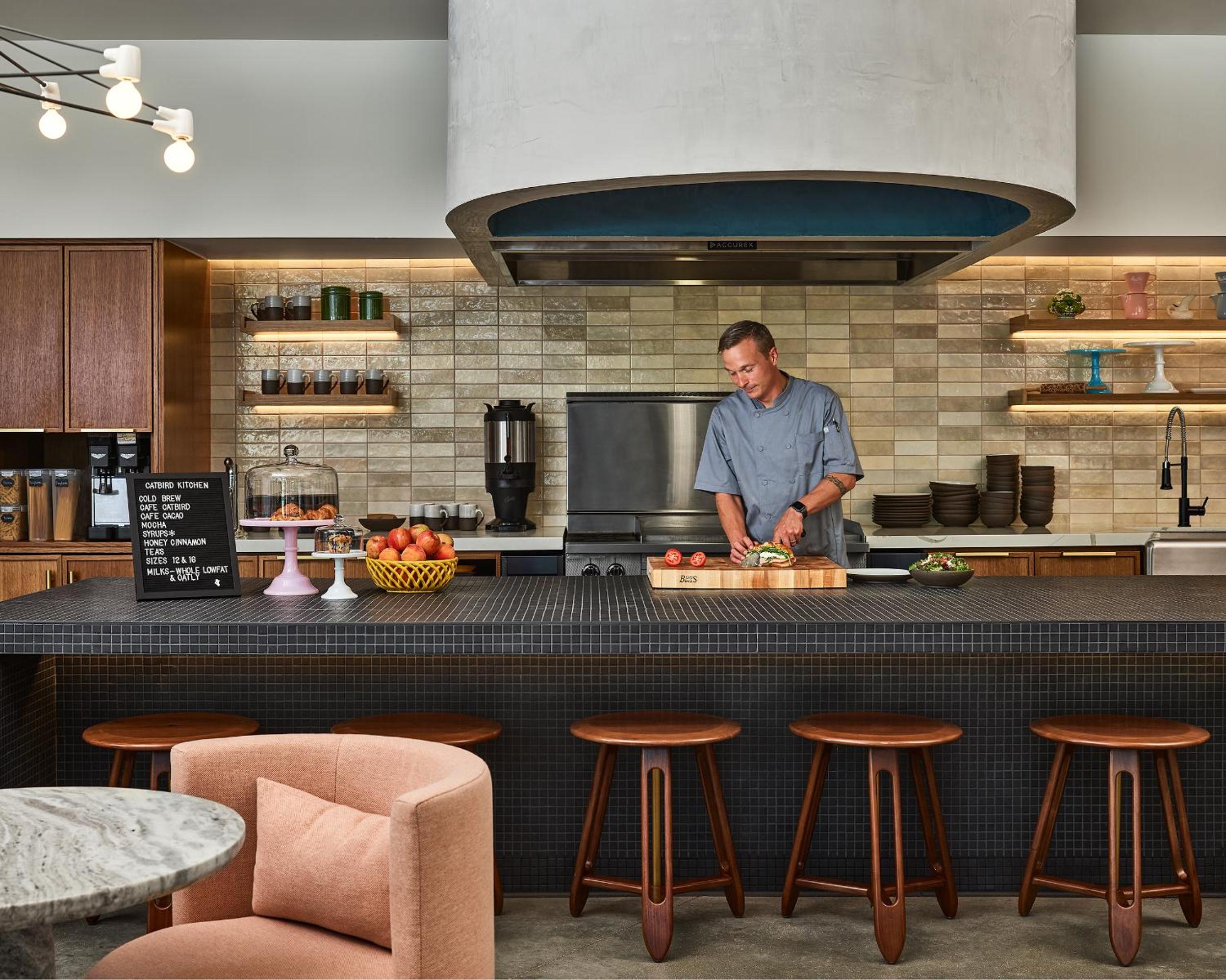 A chef prepares food behind a dark tiled kitchen counter beneath a large curved vent hood and warm tiled backsplash.