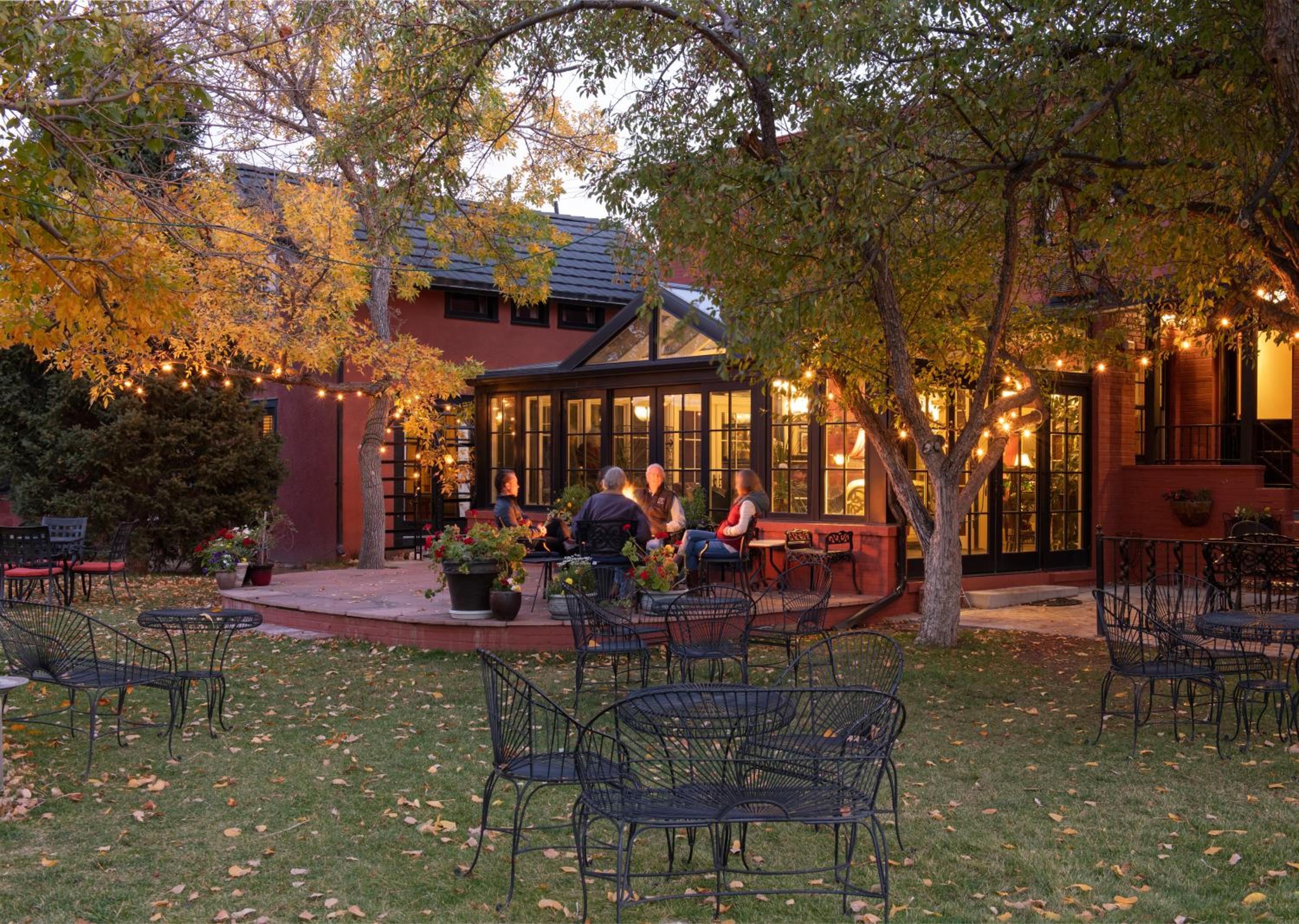 Guests sit around a fire pit on a patio outside a glass conservatory surrounded by trees lit with string lights.