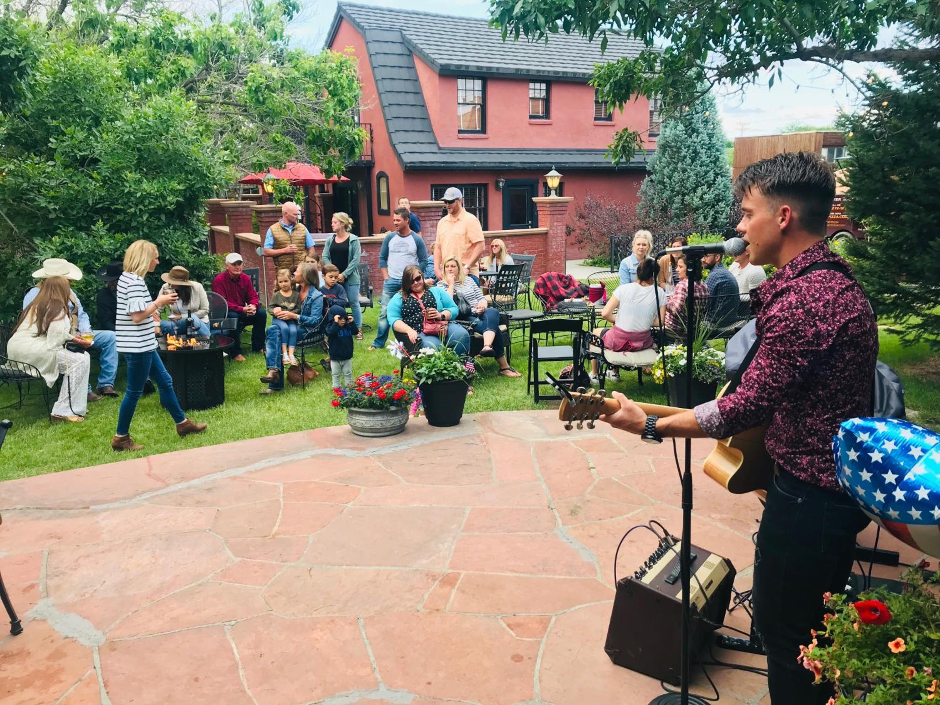 A guitarist performs on a stone terrace for an audience seated on lawn chairs in a garden.