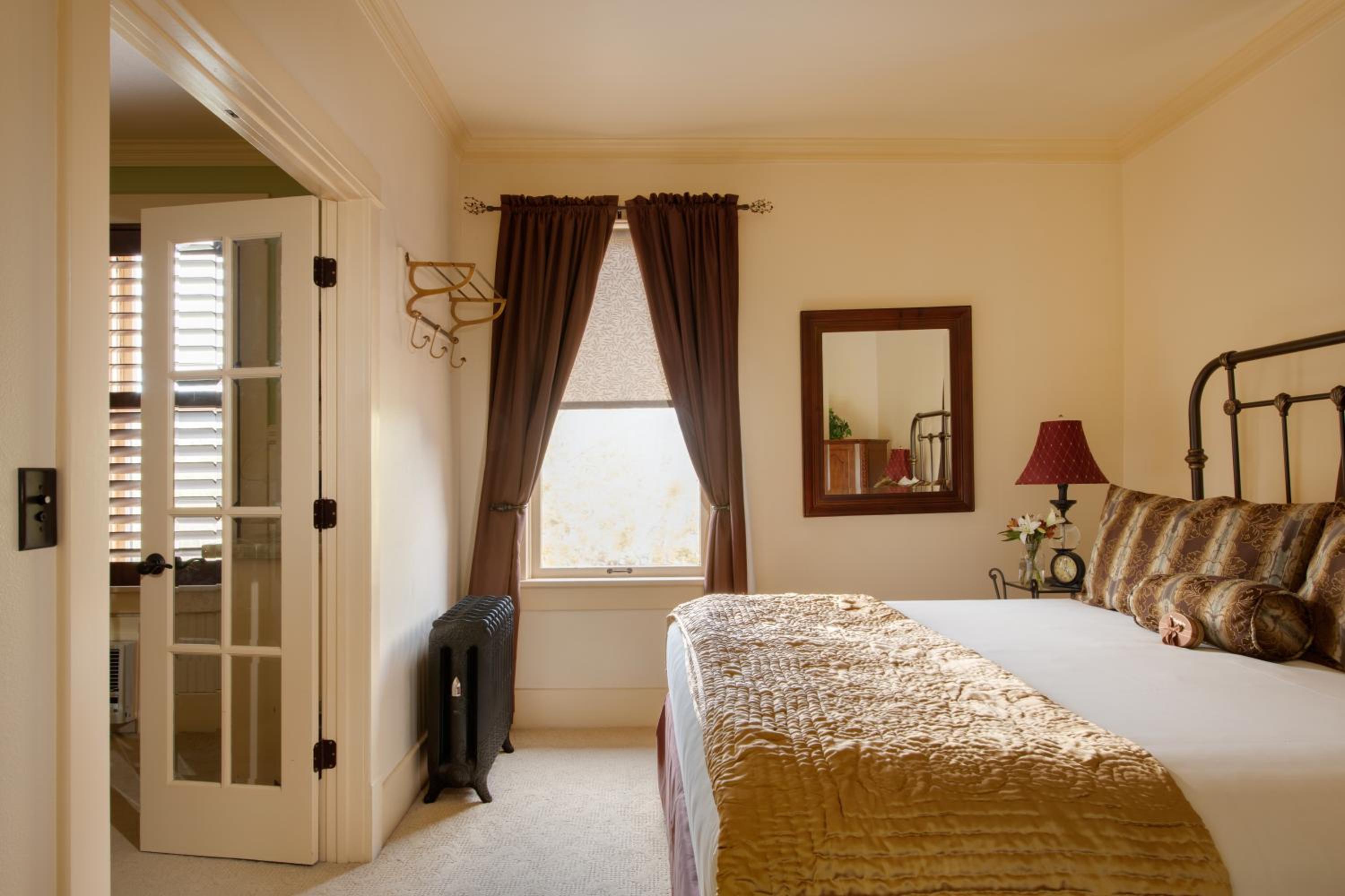 A bedroom features a wrought-iron bed with a gold quilt, dark curtains, and an open door to an adjoining room.