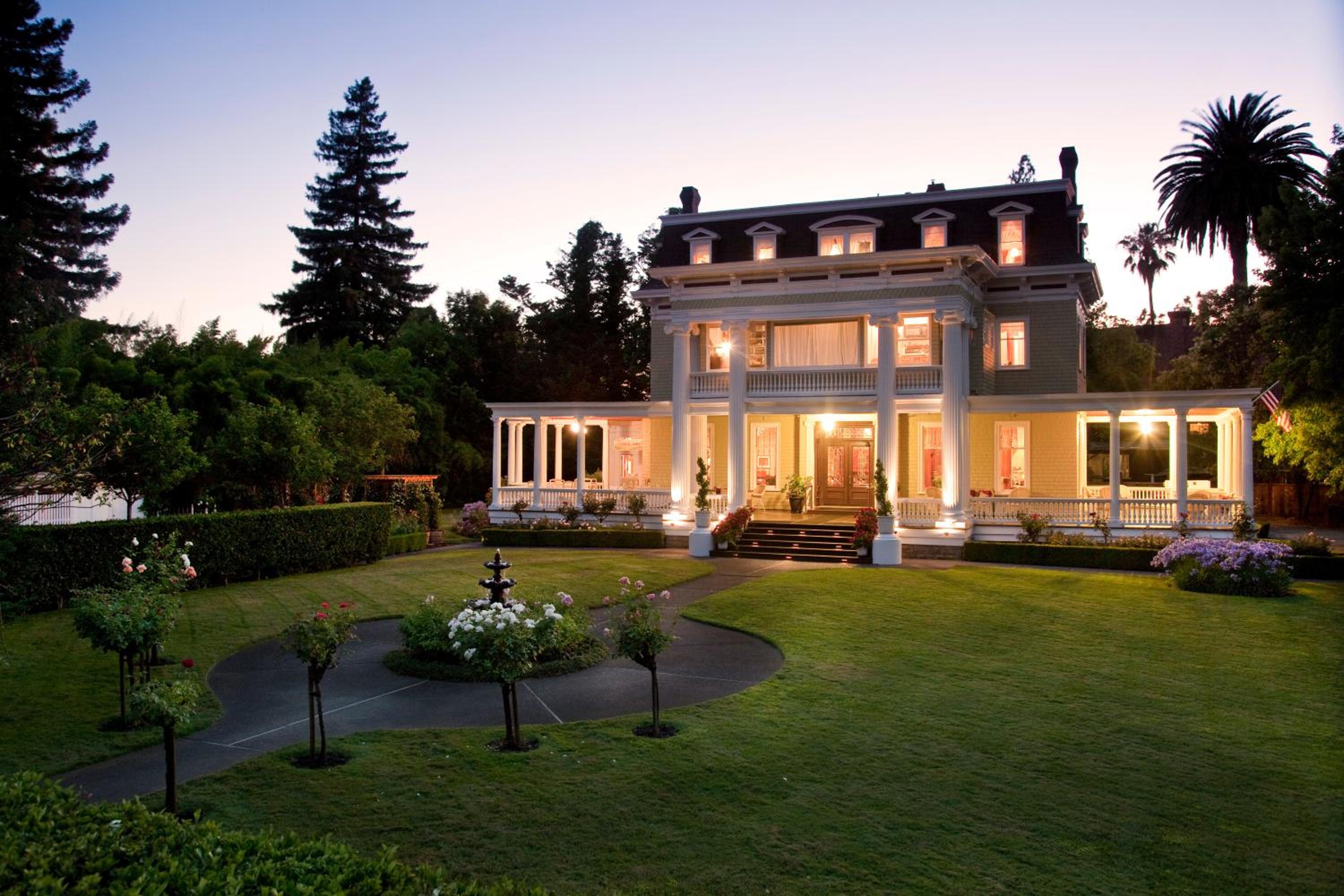 A twilight view of Churchill Manor's columned mansion with illuminated windows and a green lawn with a fountain.
