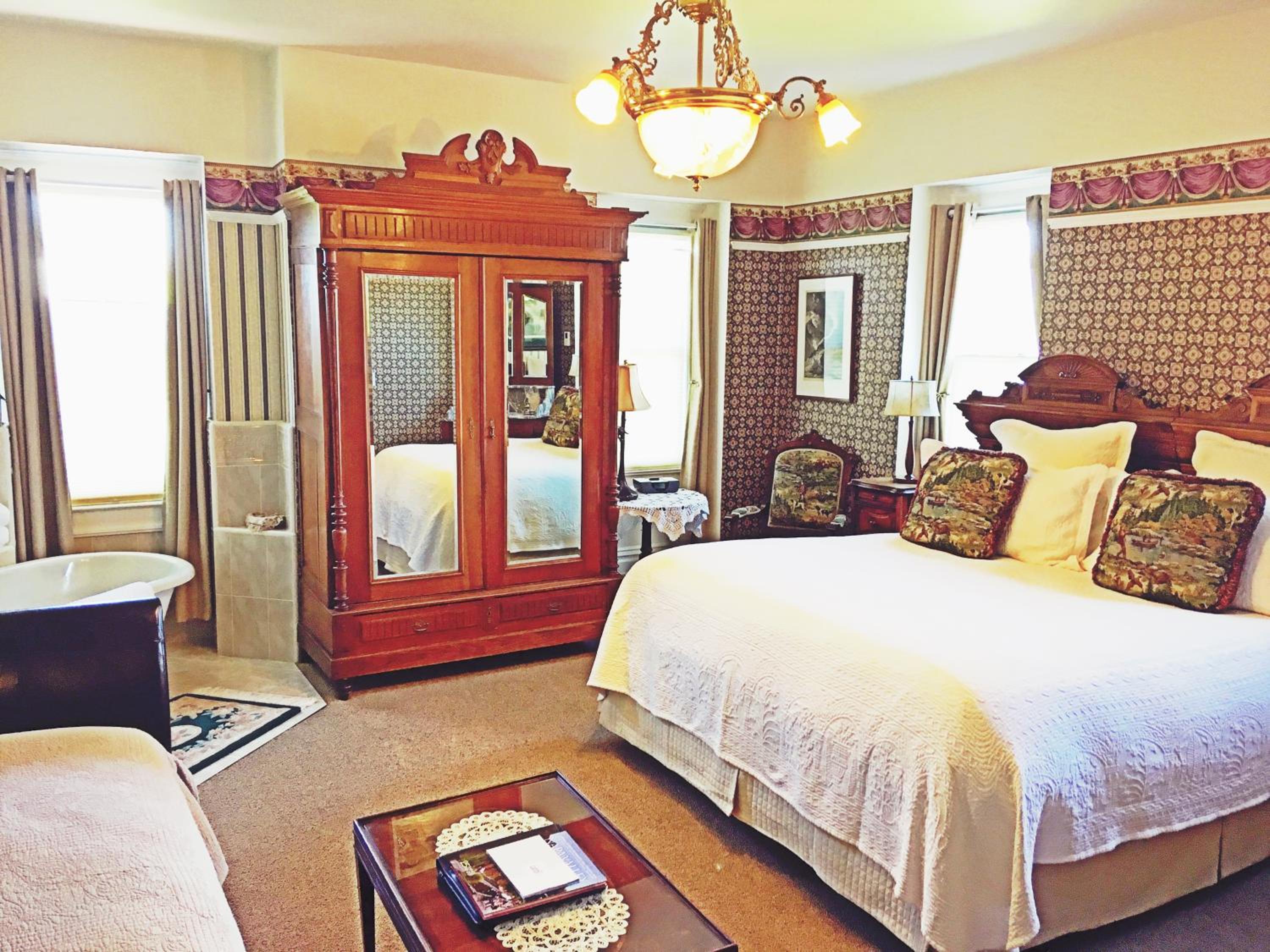 A bedroom with patterned wallpaper, a mirrored wooden armoire, and a white bed under an ornate chandelier.