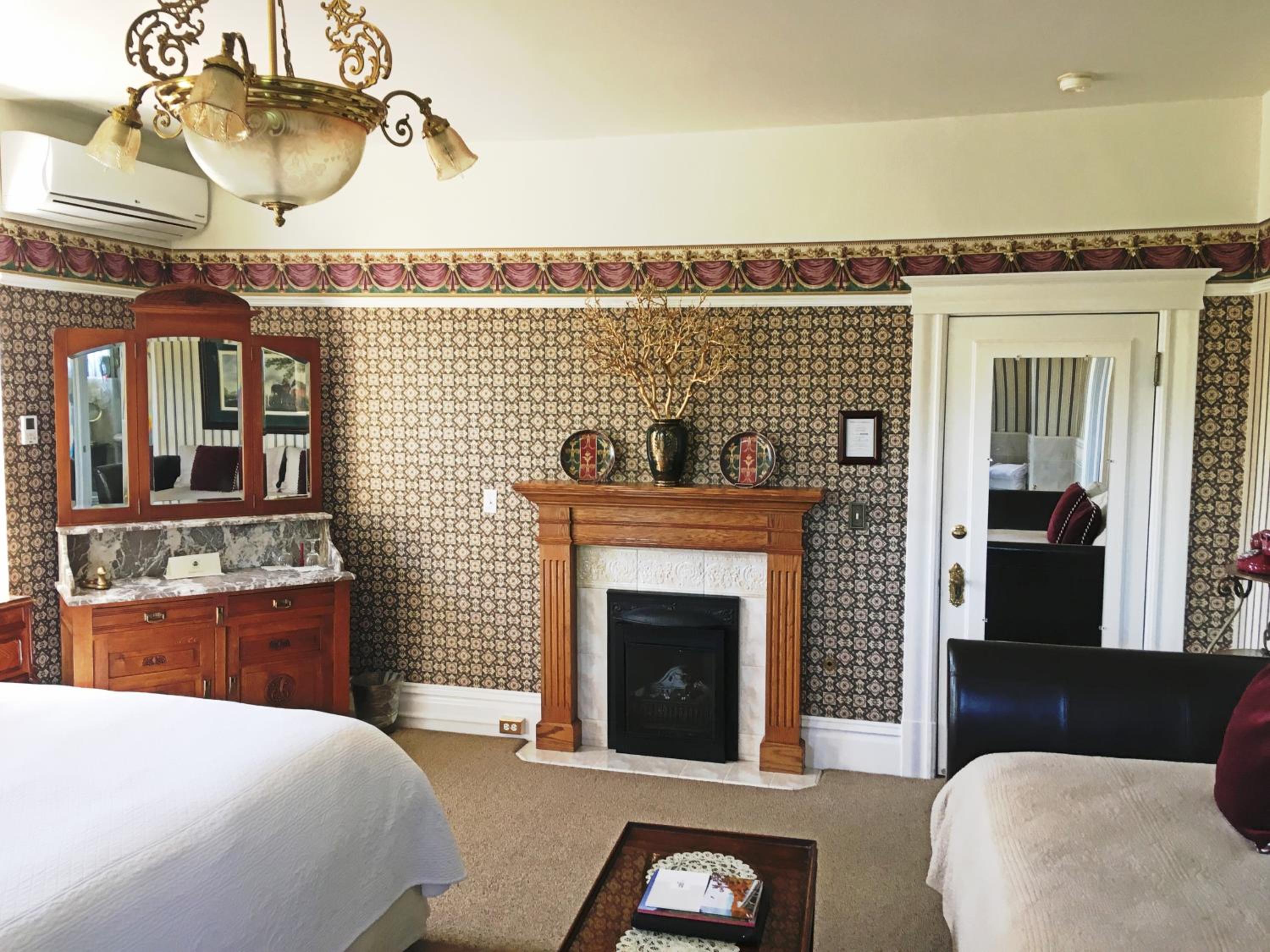 A bedroom with a wooden fireplace mantel, decorative wallpaper, and an antique dressing table with mirrors.