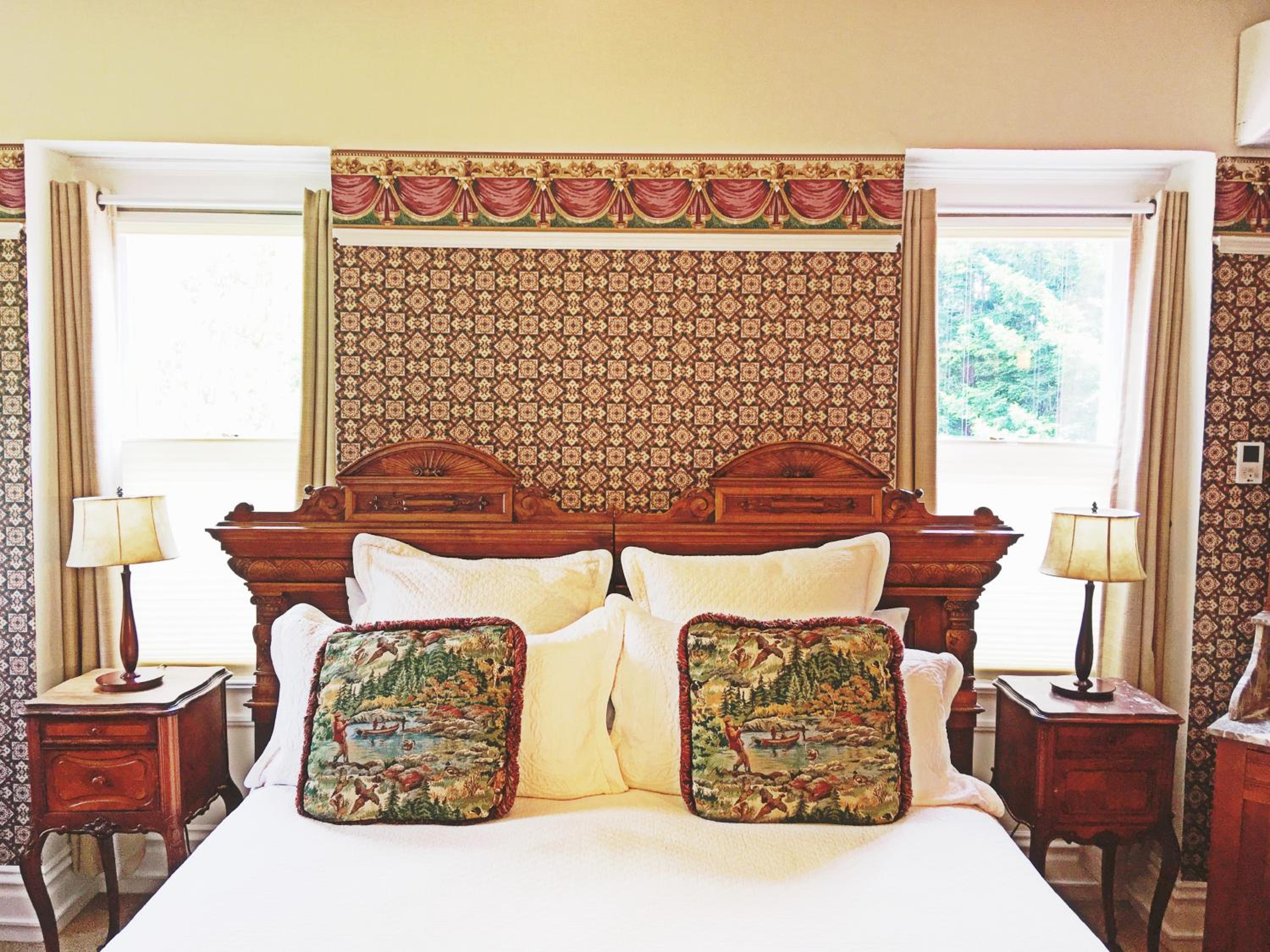 A carved wooden headboard against patterned wallpaper between curtained windows with bedside tables and lamps.
